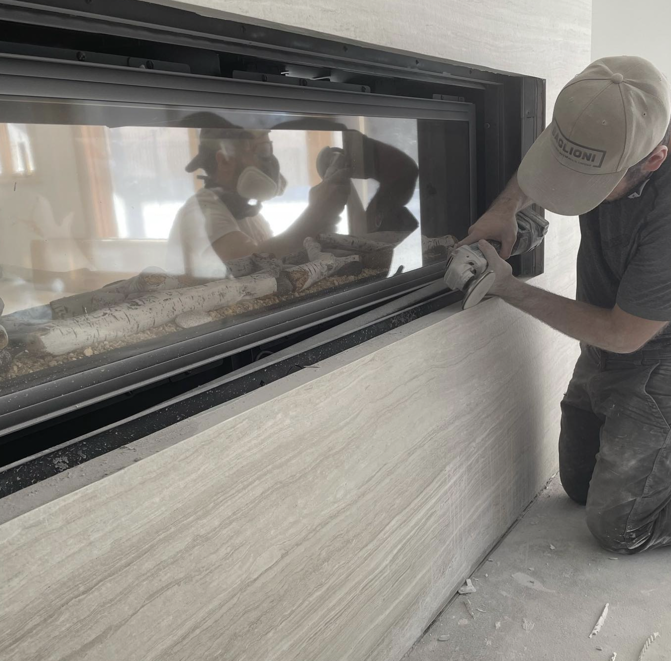A worker cutting a stone panel with a power saw, reflected in a glass window, while kneeling on the floor.