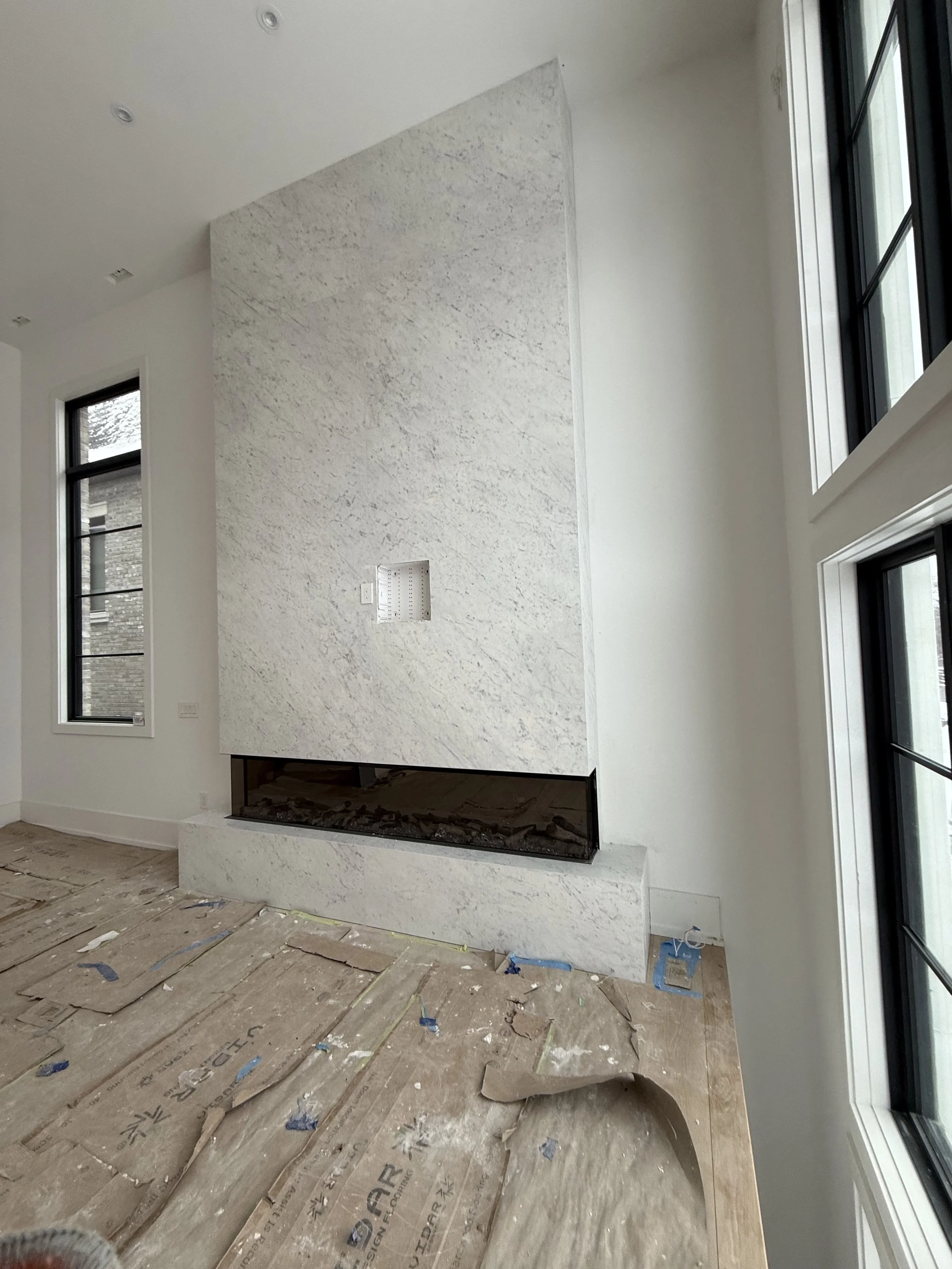 Interior of a room under construction with a modern fireplace featuring a white marble surround, multiple large windows, and a wooden floor with protective covering.