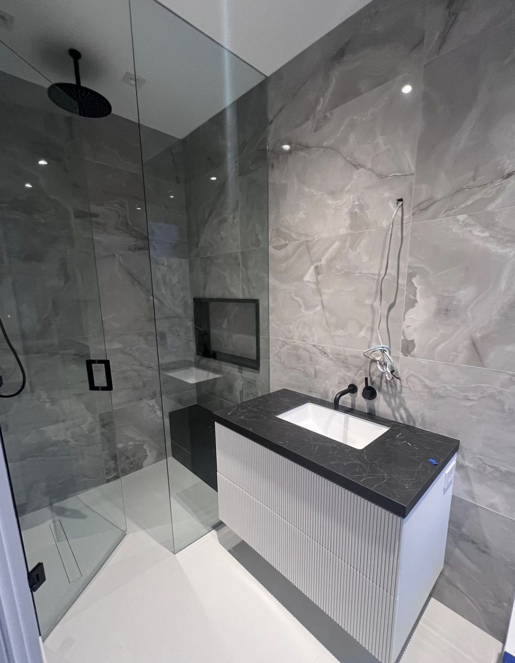 Modern bathroom with a glass-enclosed shower, black wall-mounted shower head, gray marble-tiled walls, and a black granite countertop with an integrated sink and textured white cabinet.