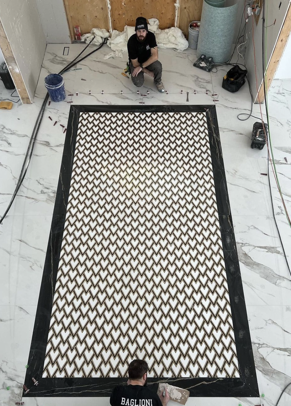 Two workers laying patterned tiles inside a room with marble floors, one kneeling at the bottom and one crouching at the top, with tools and construction materials around.