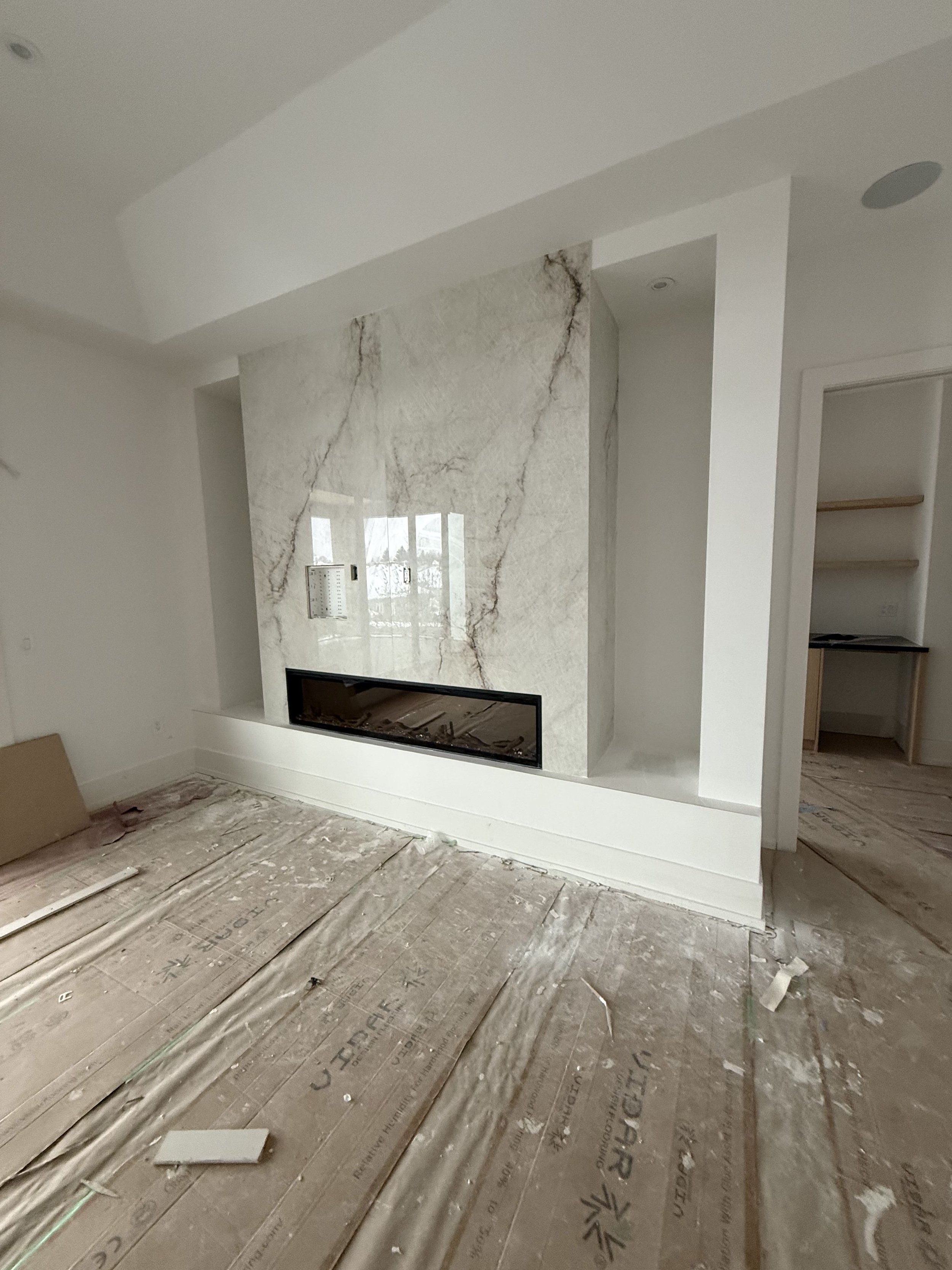 Modern living room under construction with white walls, a marble accent wall with a built-in fireplace, and unfinished flooring.