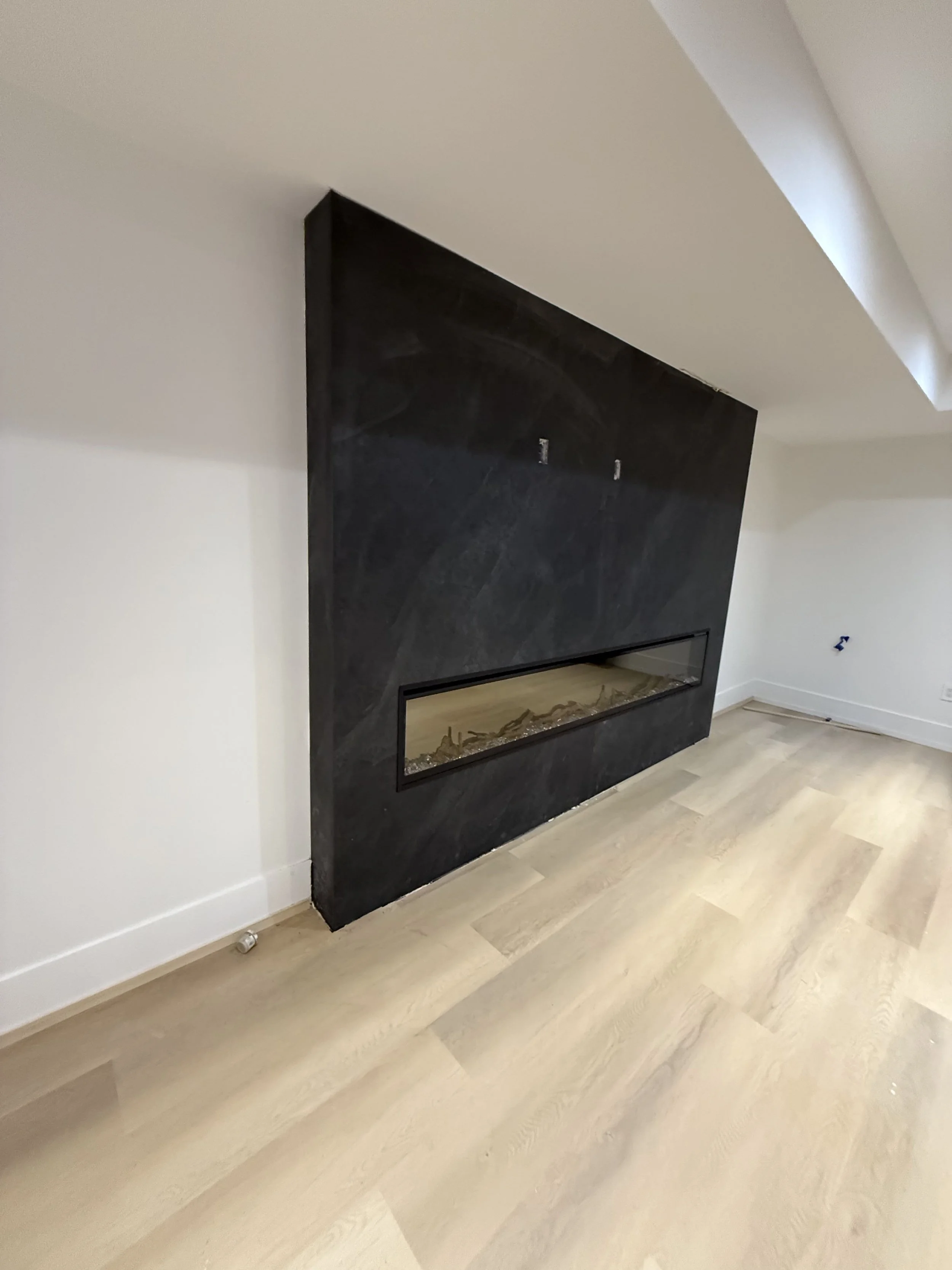 Empty living room with a black modern fireplace built into a dark wall panel, light wooden flooring, and plain white walls.