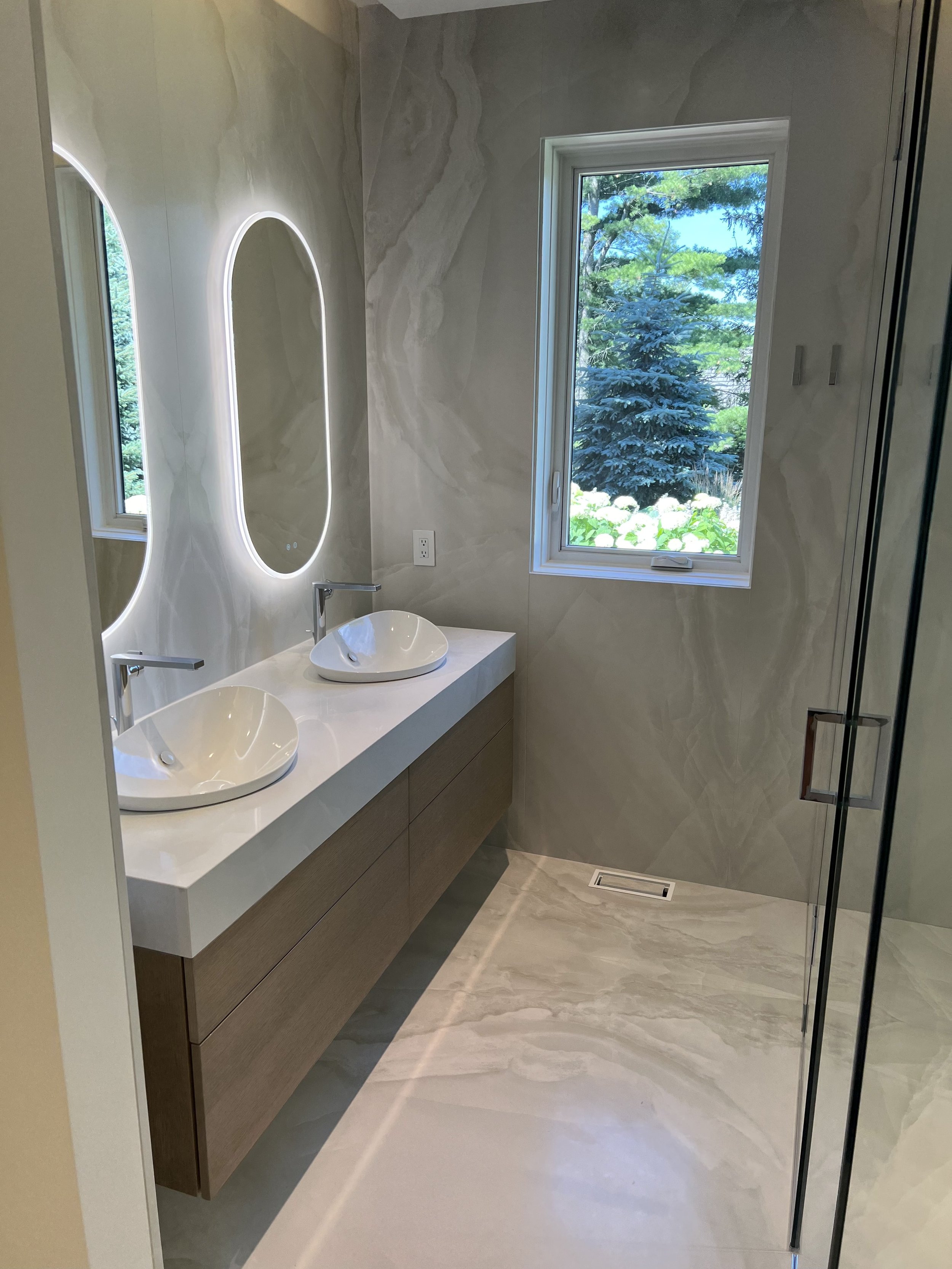 Modern bathroom with a double vanity featuring vessel sinks and backlit mirrors, beige marbled walls and floors, a large window showing greenery outside, and a glass shower door.