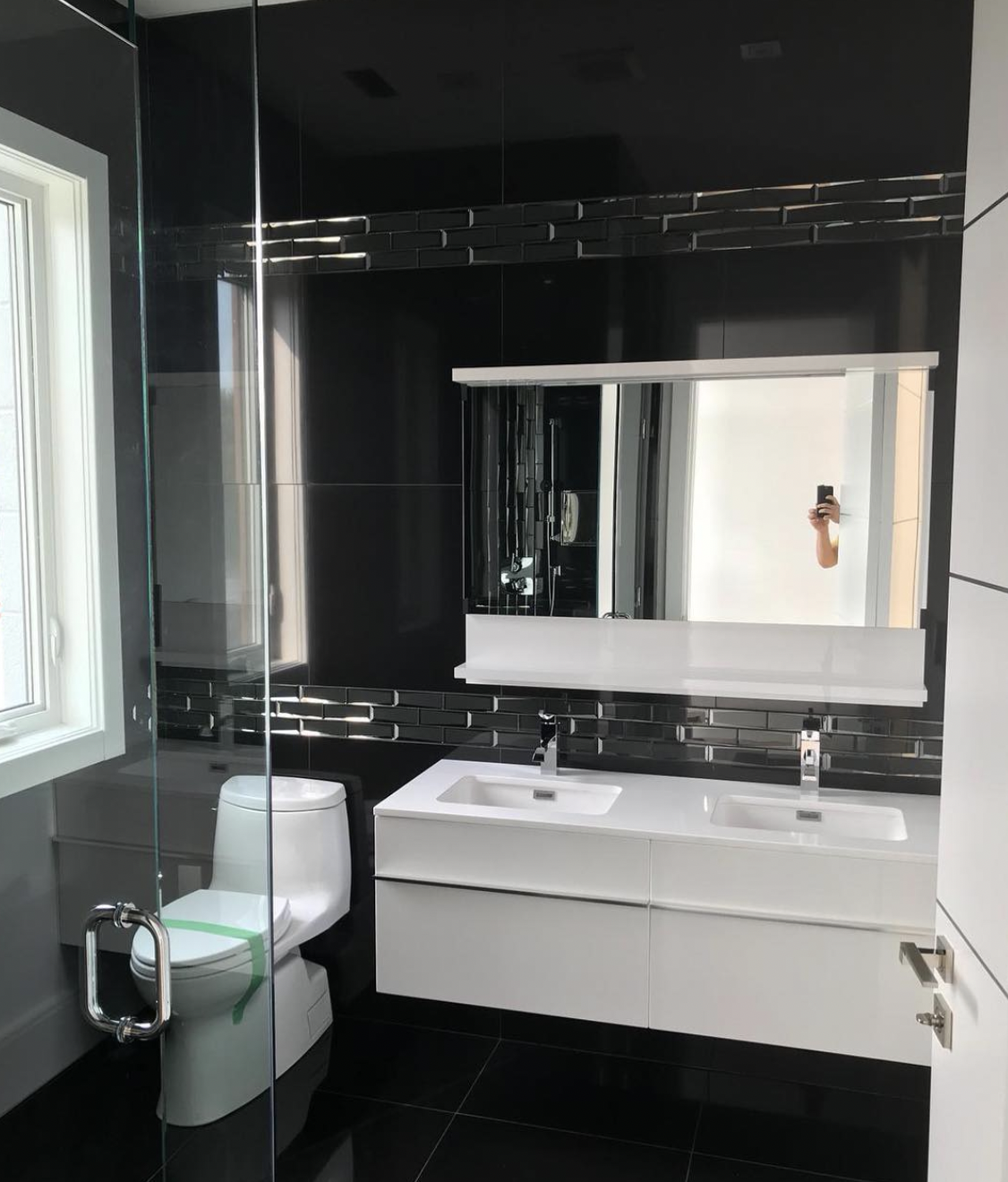 Modern bathroom with black tiled walls and floor, a white double sink vanity with a large mirror, and a toilet. A person is taking a photo of the mirror with a smartphone in their hand.