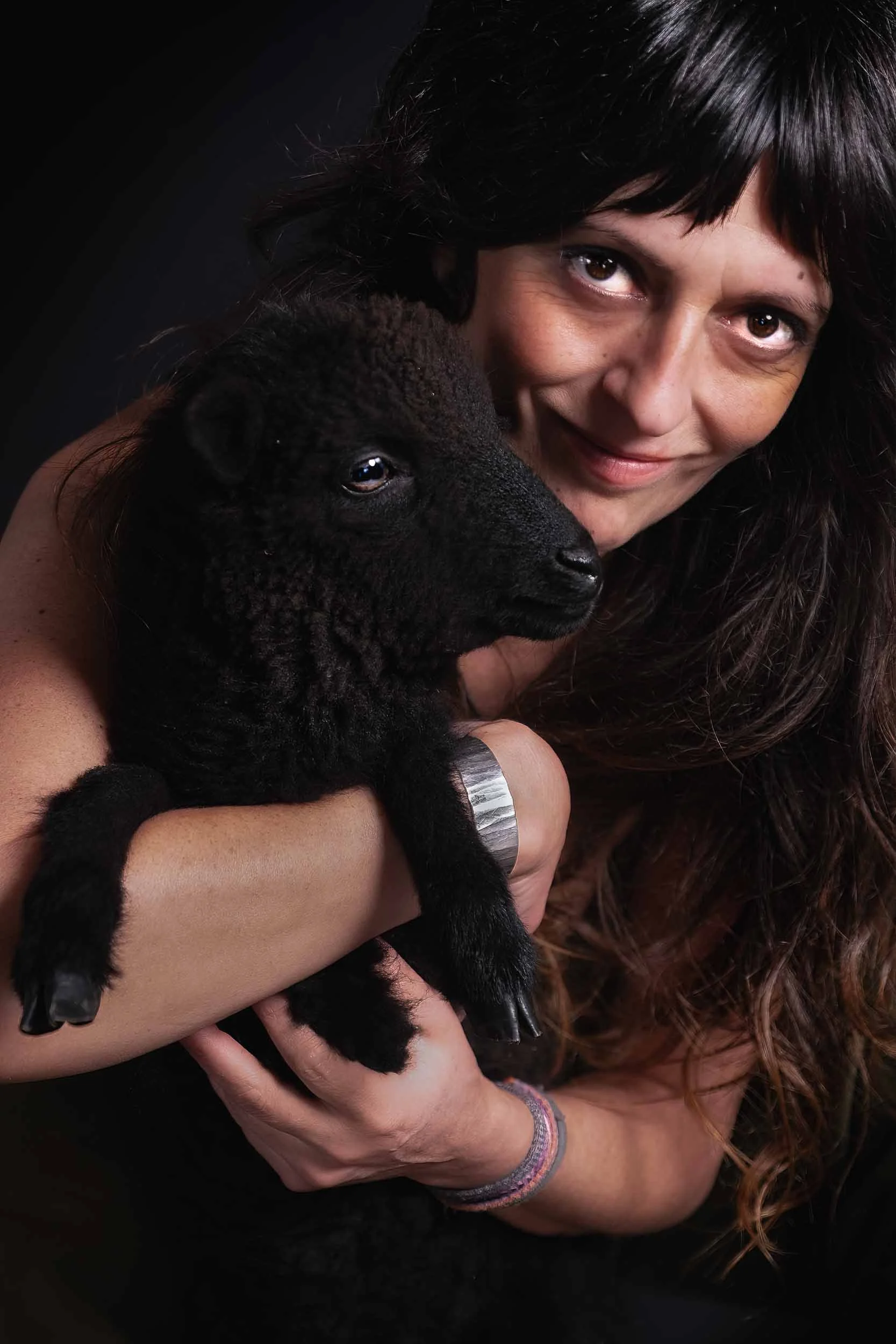 Primer plano de una joven con un cordero negro en brazos, estilo personal rústico con enfoque en la mirada y texturas naturales.
