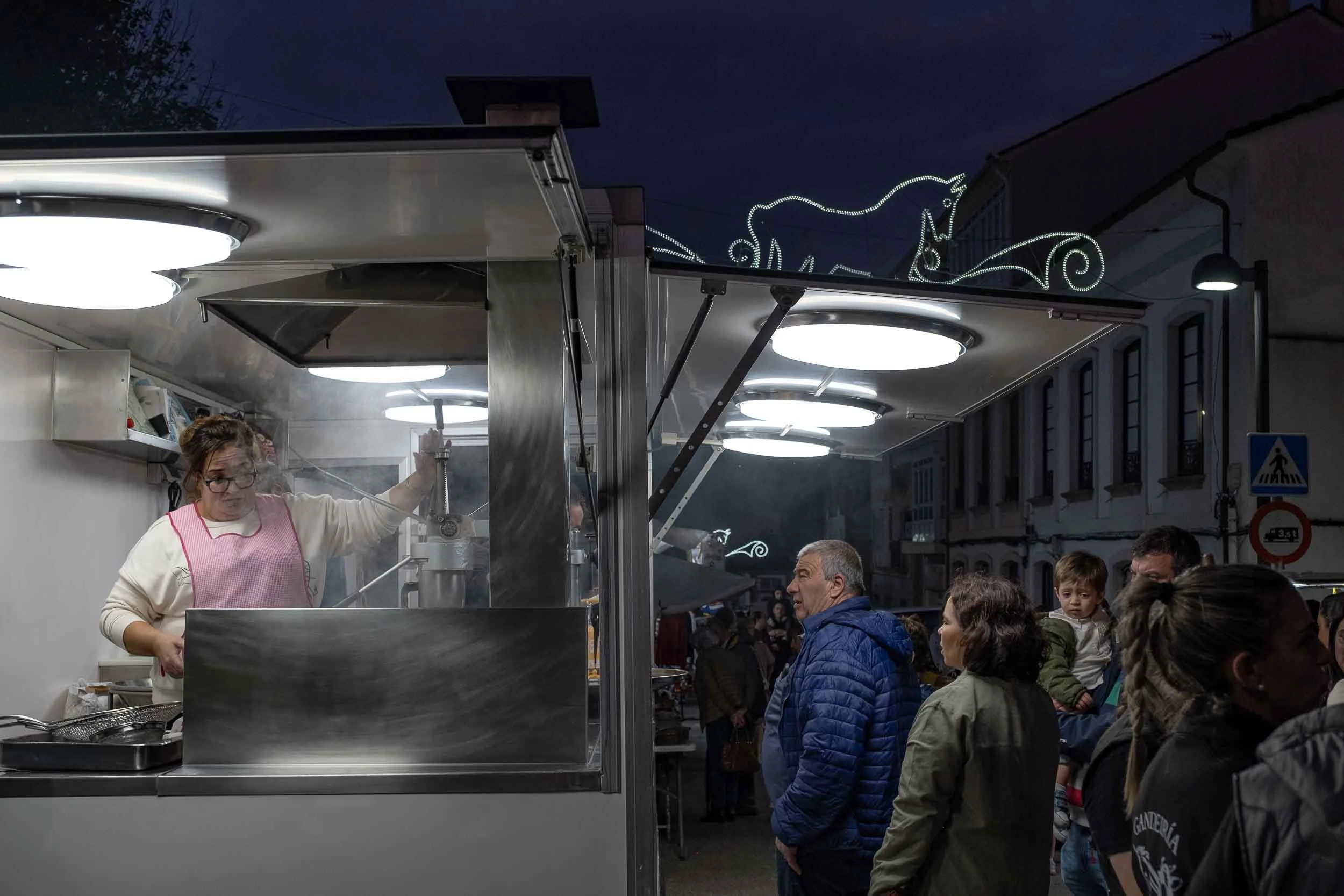 Fotografía nocturna de un puesto de churrería iluminado en el ferial de Mondoñedo. Captura la atmósfera festiva y tradicional de las noches en As San Lucas.