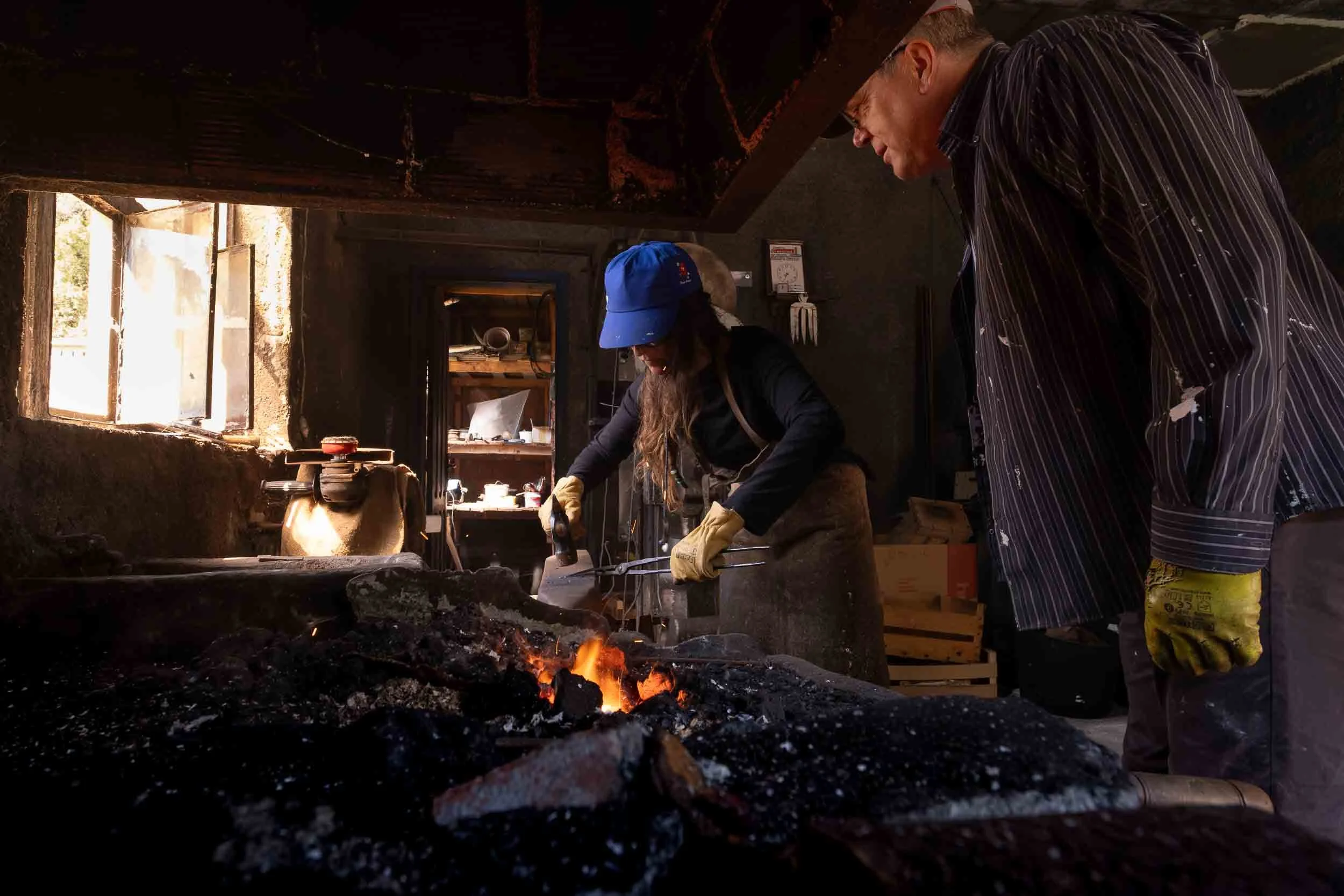 Maestro ferreiro supervisando a una aprendiz mientras trabaja el metal en una fragua de carbón.
