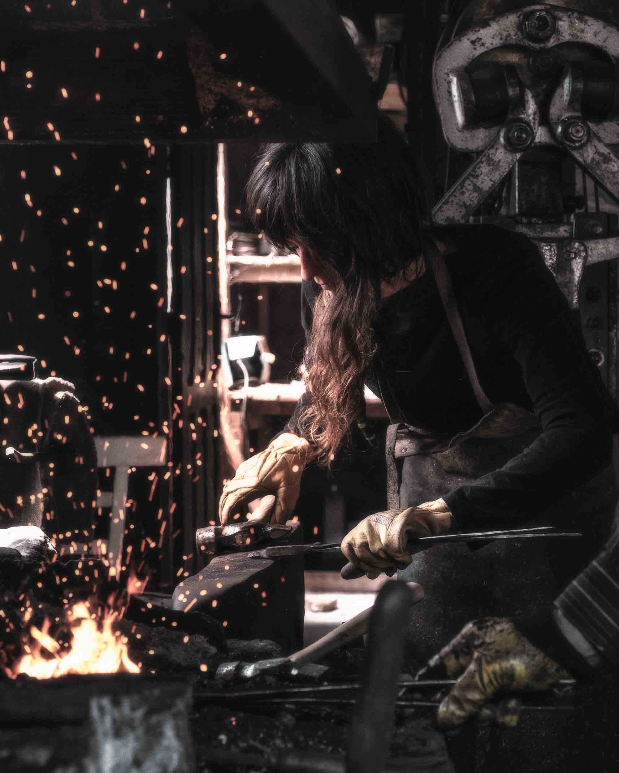 Un primer plano de la artesana golpeteando el hierro candente en la forja. La iluminación dramática resalta la textura del metal y la concentración.