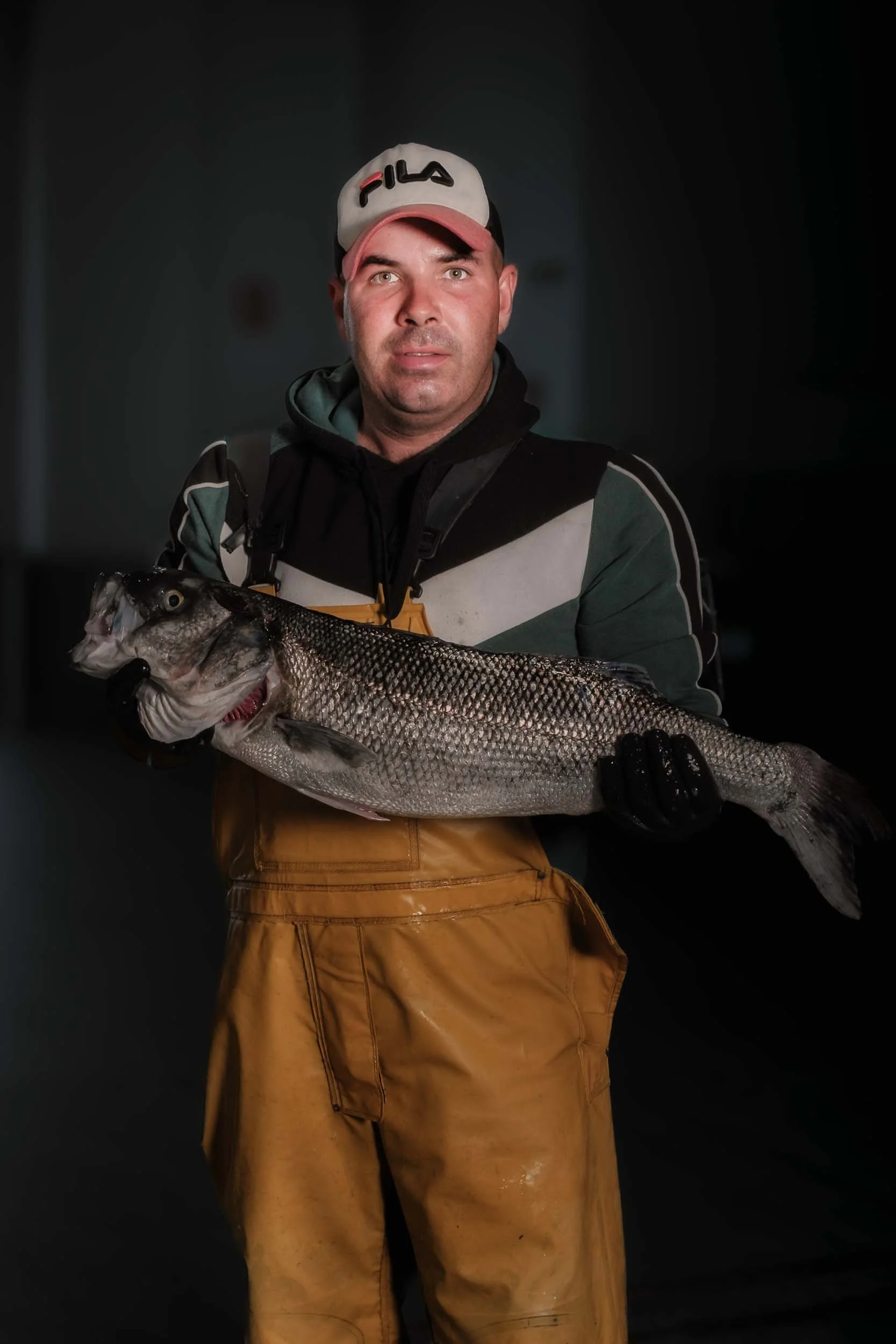 Pescador local mostrando con orgullo una lubina de gran tamaño, iluminación dramática en entorno profesional de pesca.