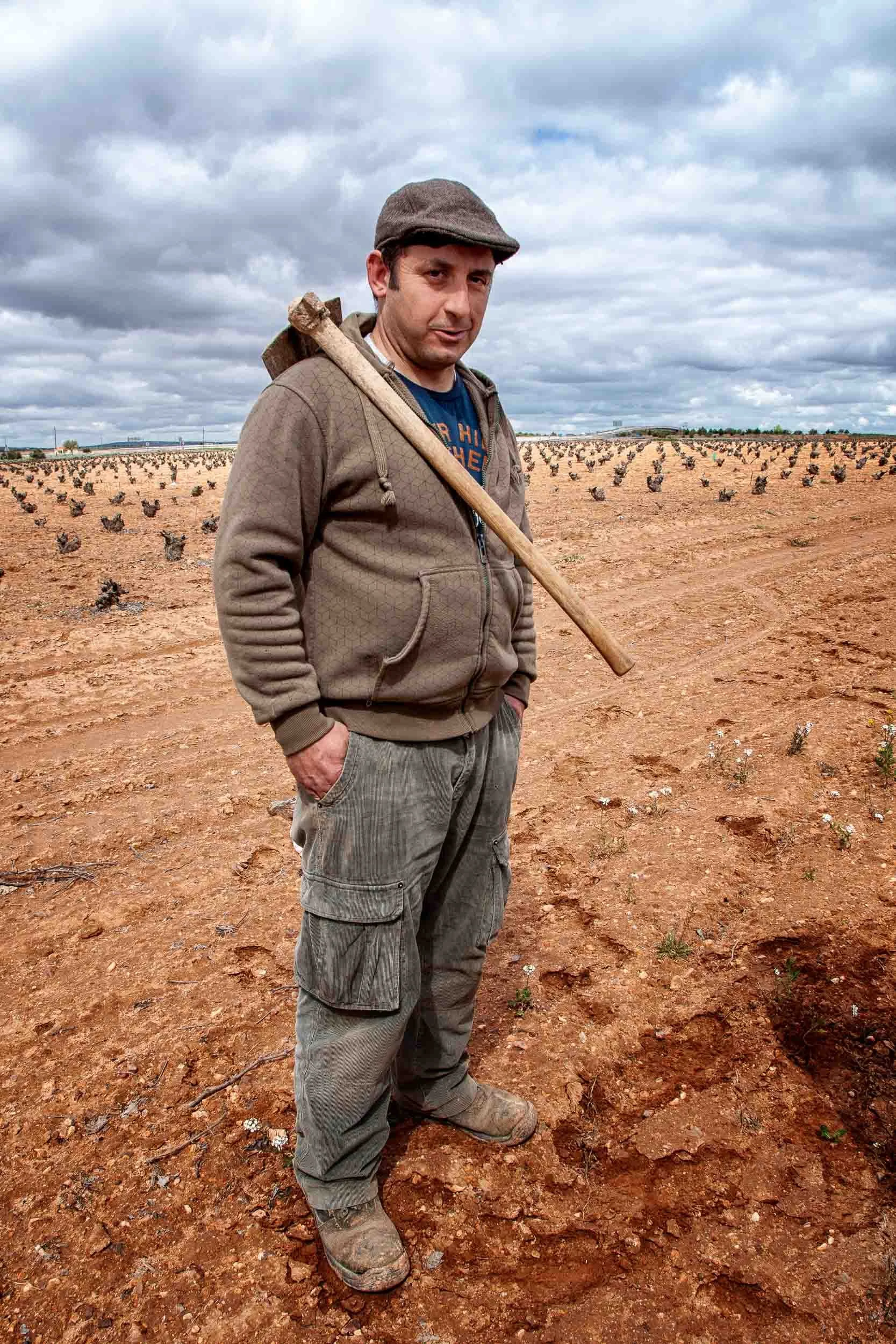 Agricultor descansando entre hileras de viñedos bajo un cielo nublado, retrato ambiental de la cultura del vino.