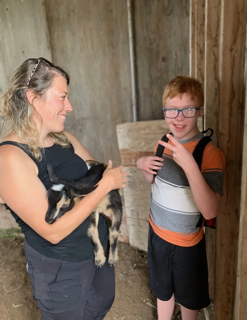 A woman is holding a goat smiling at a young man giving a peace sign.