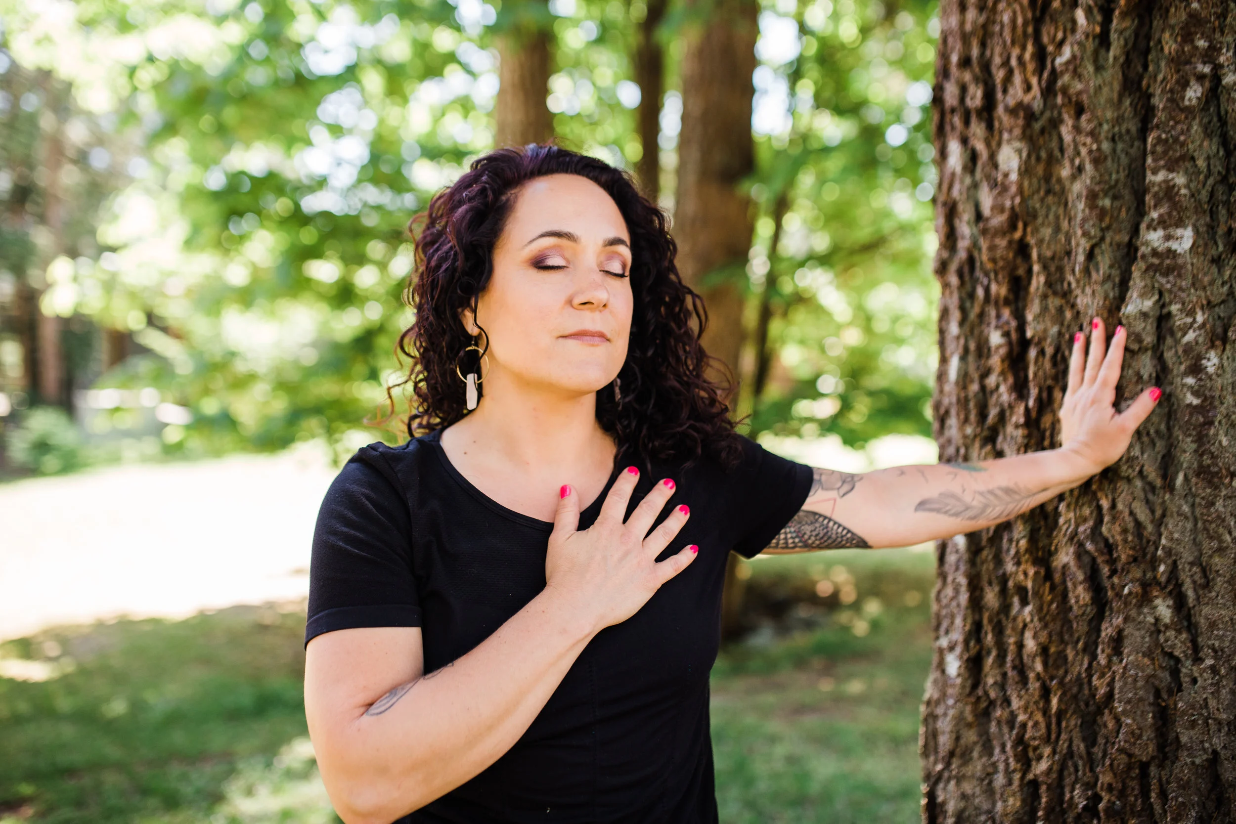 A woman with dark, curly hair and tattoos on her arm is outdoors hugging a tree with her eyes closed, wearing a black shirt and pink nail polish.