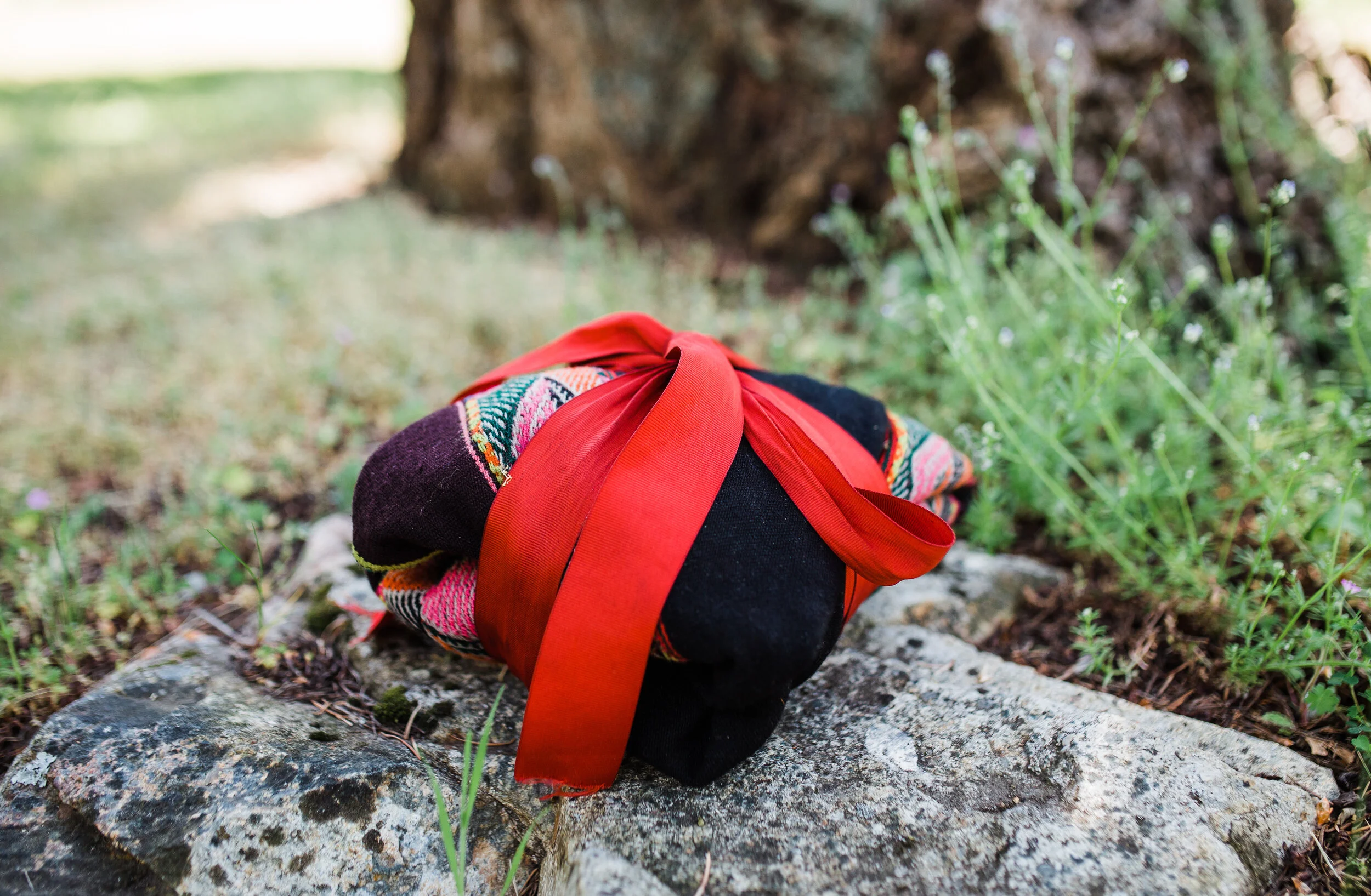 A small colorful bundle with indigenous patterns, tied with a bright red ribbon, resting on a rock outdoors.