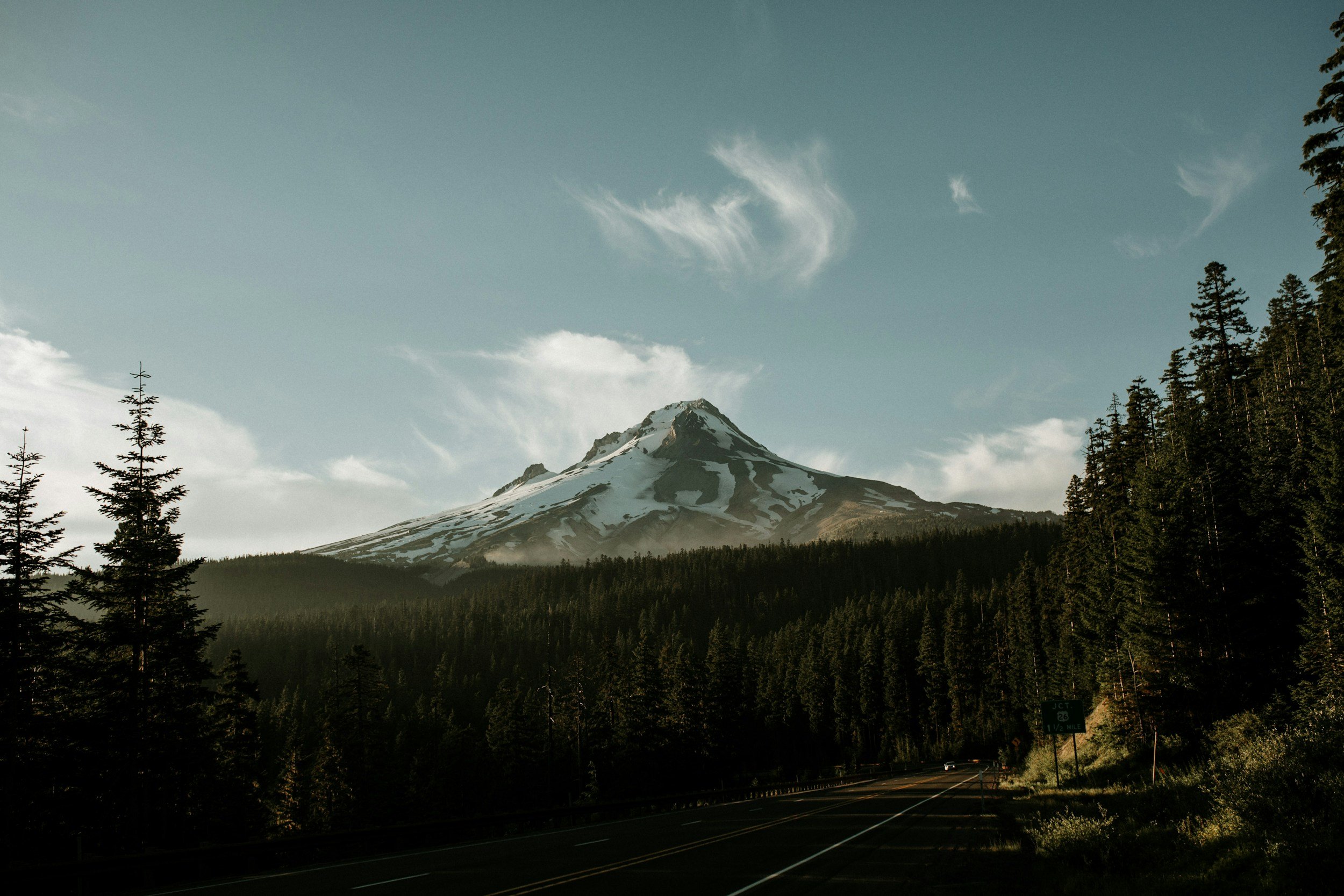 A snow-capped mountain, possibly Mount Hood, with a forest of conifer trees in the foreground and a clear sky with some clouds.
