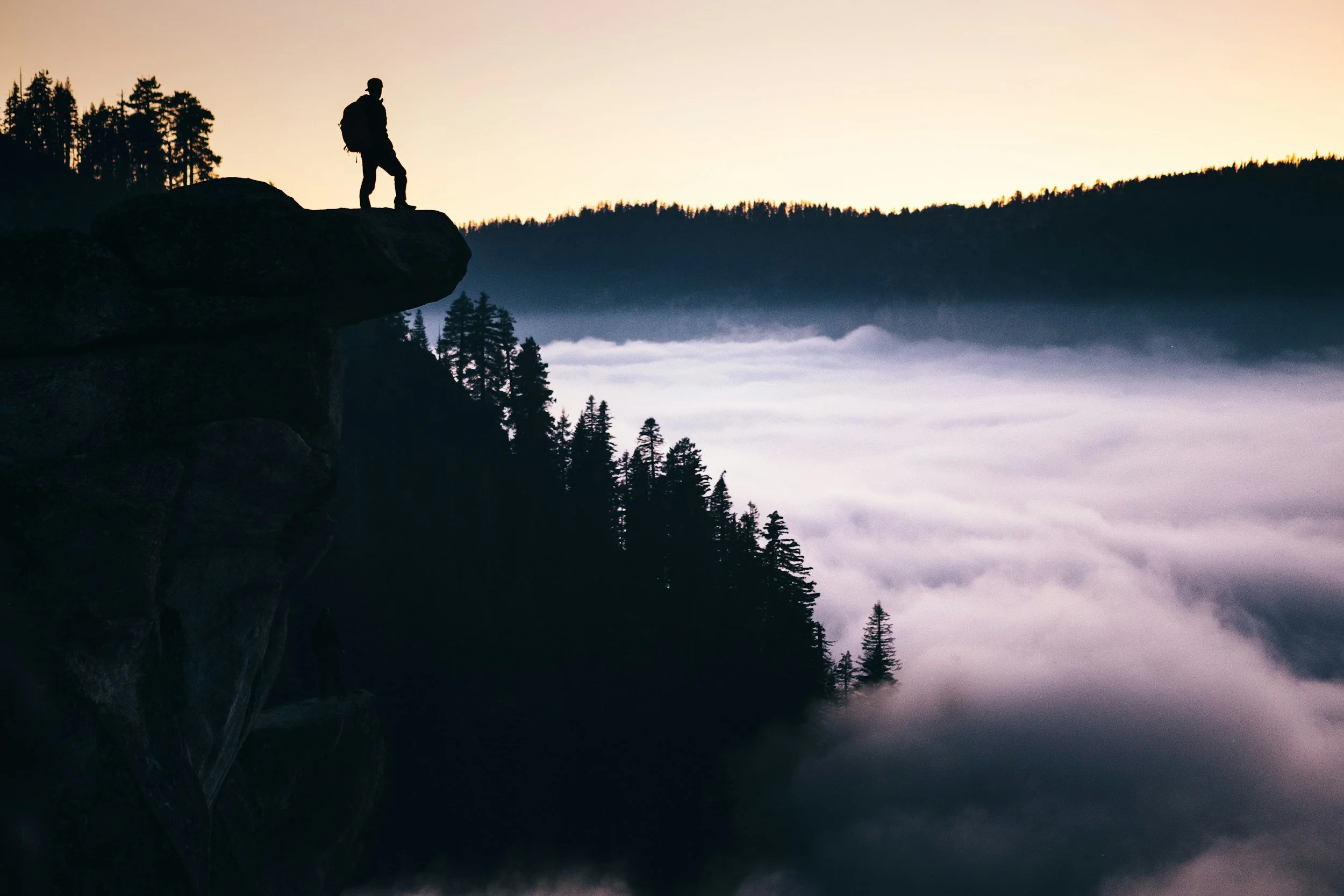 Silhouette of a hiker standing on a rocky ledge overlooking a foggy forested valley at sunrise or sunset.
