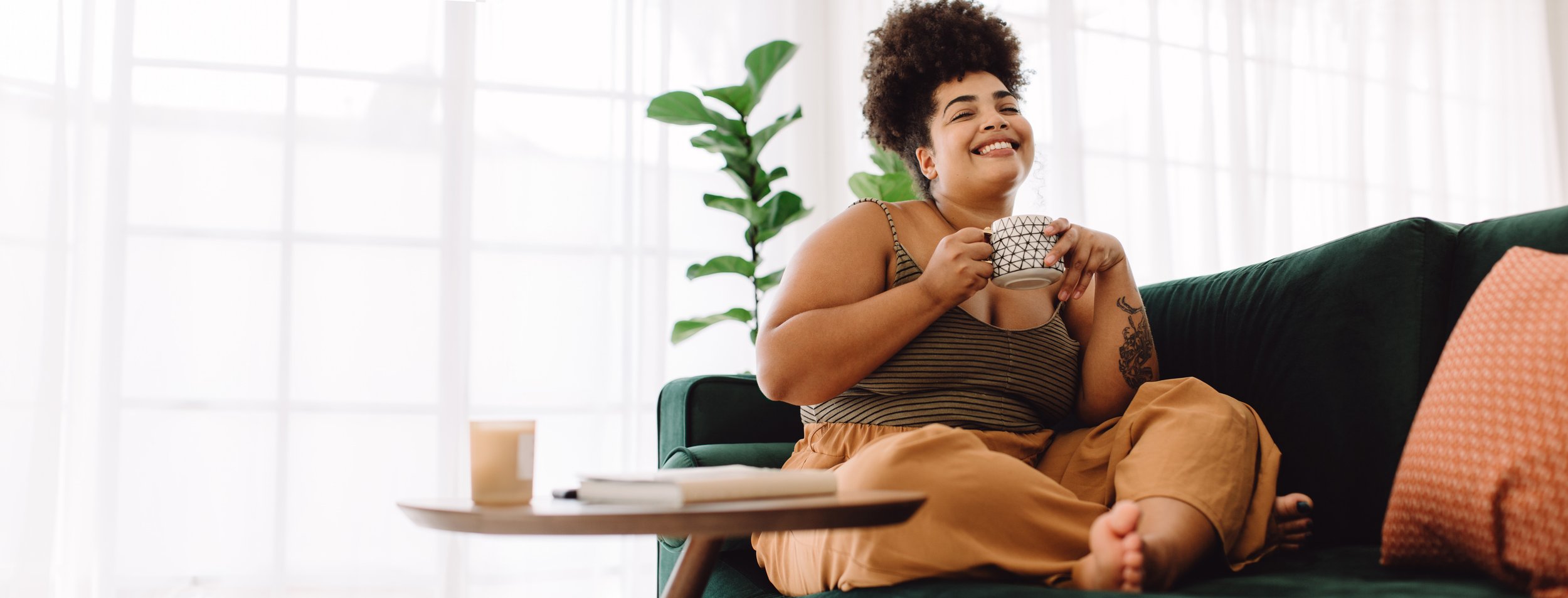 A woman with curly hair and a tattoo on her arm sitting on a green couch, smiling while holding a patterned mug. She is wearing a striped tank top and loose pants, with sunlight coming through large windows and a plant in the background.