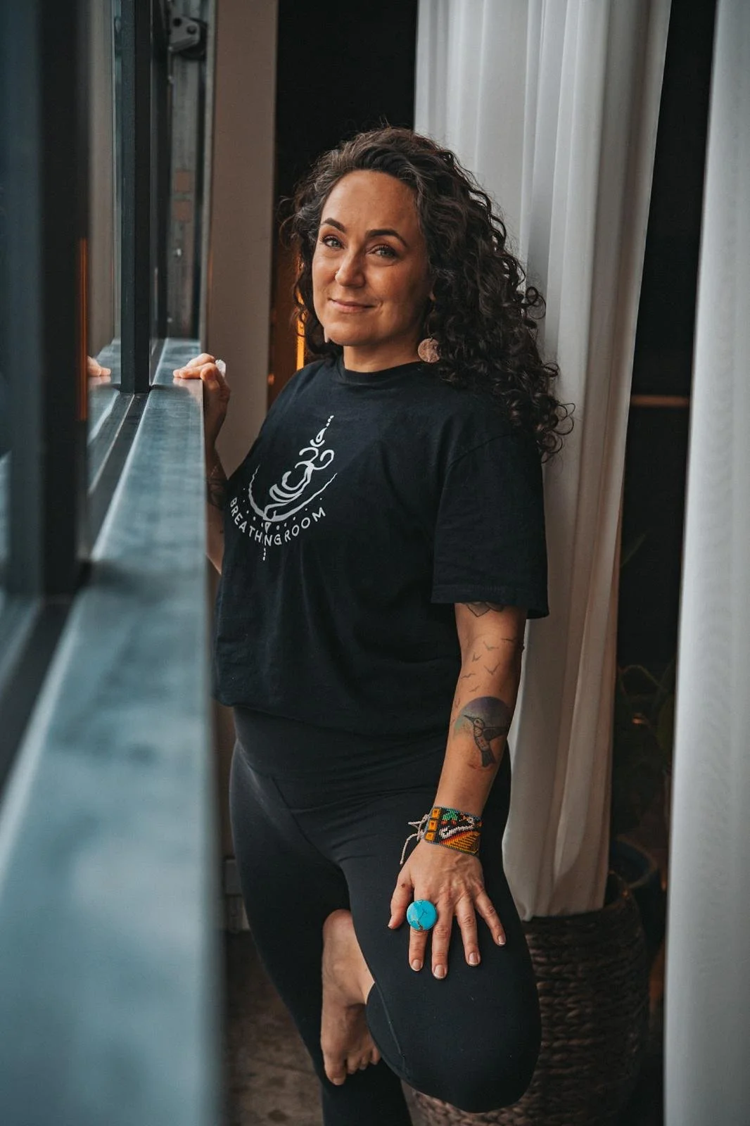 Woman with curly hair and tattoos standing near a window, wearing a black t-shirt with a breathing room logo, black pants, a turquoise ring, and bracelets.
