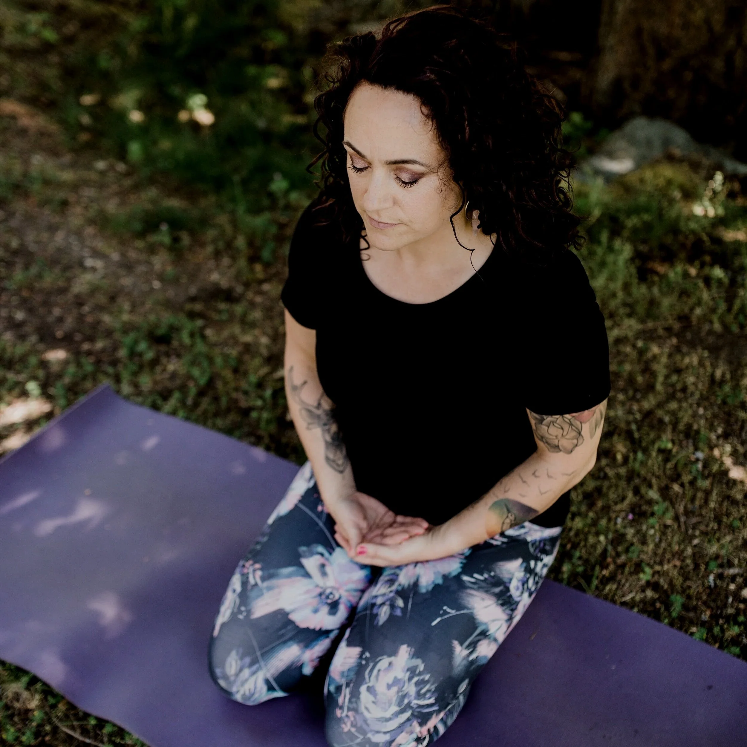 A woman with dark, curly hair and tattoos on her arms is practicing yoga outdoors on a purple mat, with greenery and dirt surrounding her.