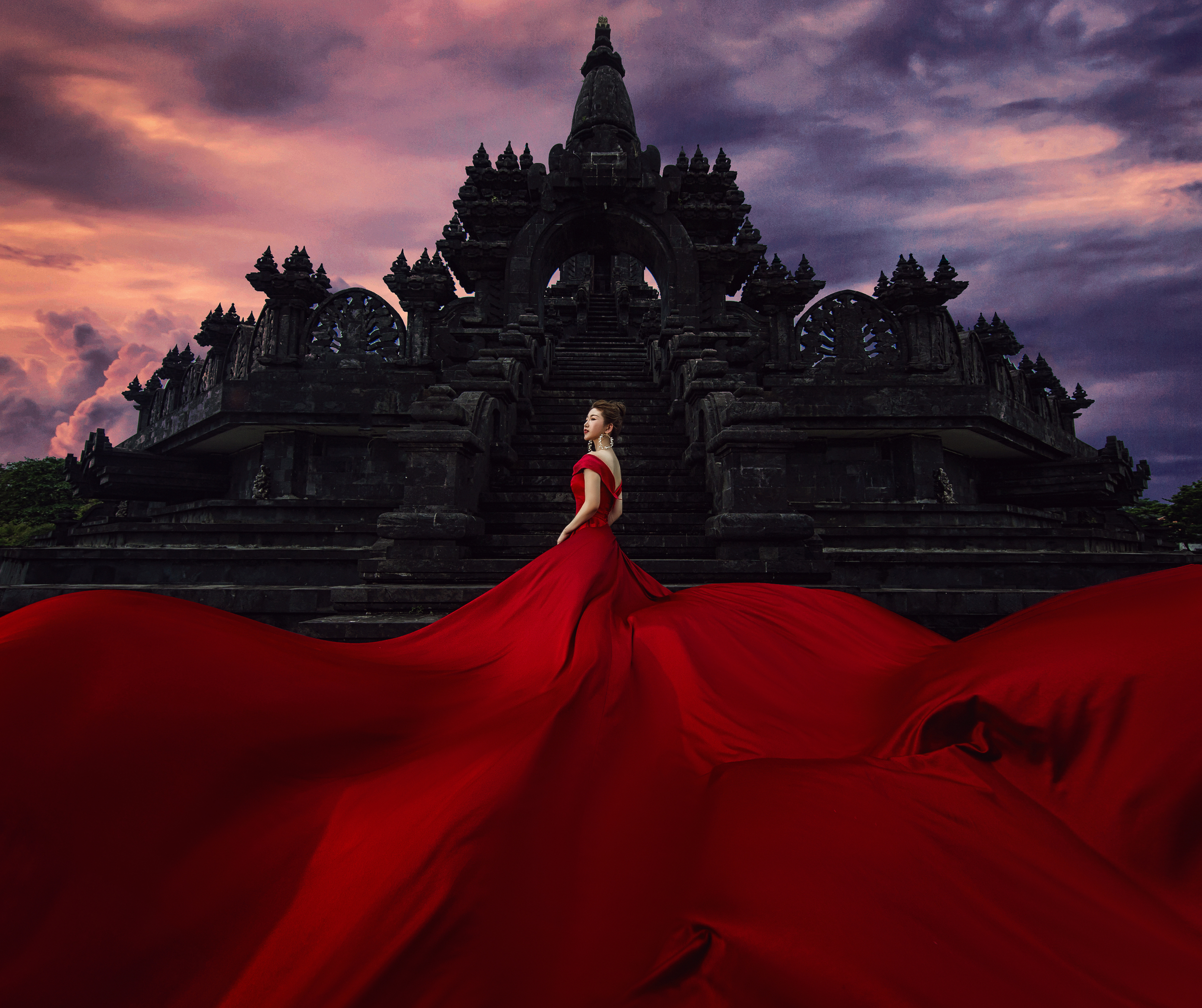 Woman in red dress with long train standing on the steps of an ancient dark stone temple, with a pink and purple cloudy sky in the background.