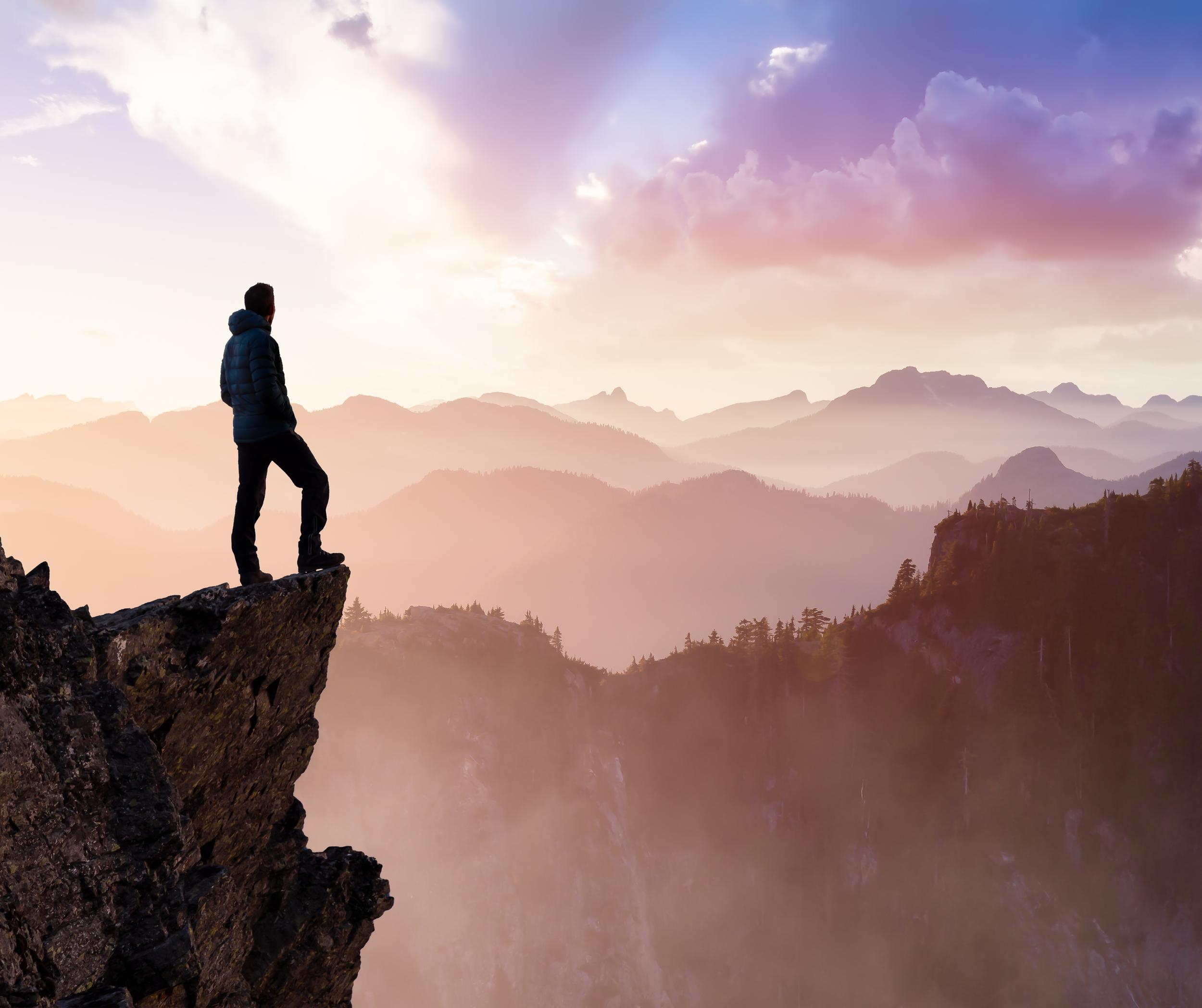 A person wearing a blue jacket standing on a rocky outcrop, gazing at a mountain landscape under a colorful sky with clouds at sunset or sunrise.