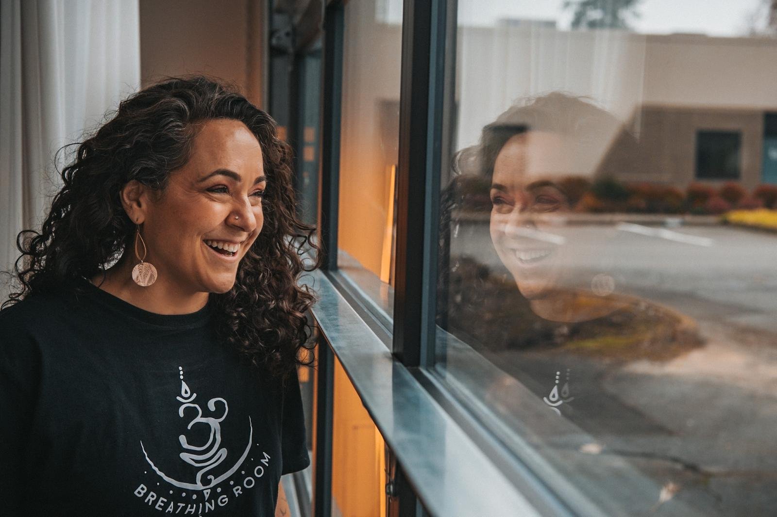 Woman with curly dark hair smiling and looking out window, with her reflection visible on the glass, indoors near a wooden structure.