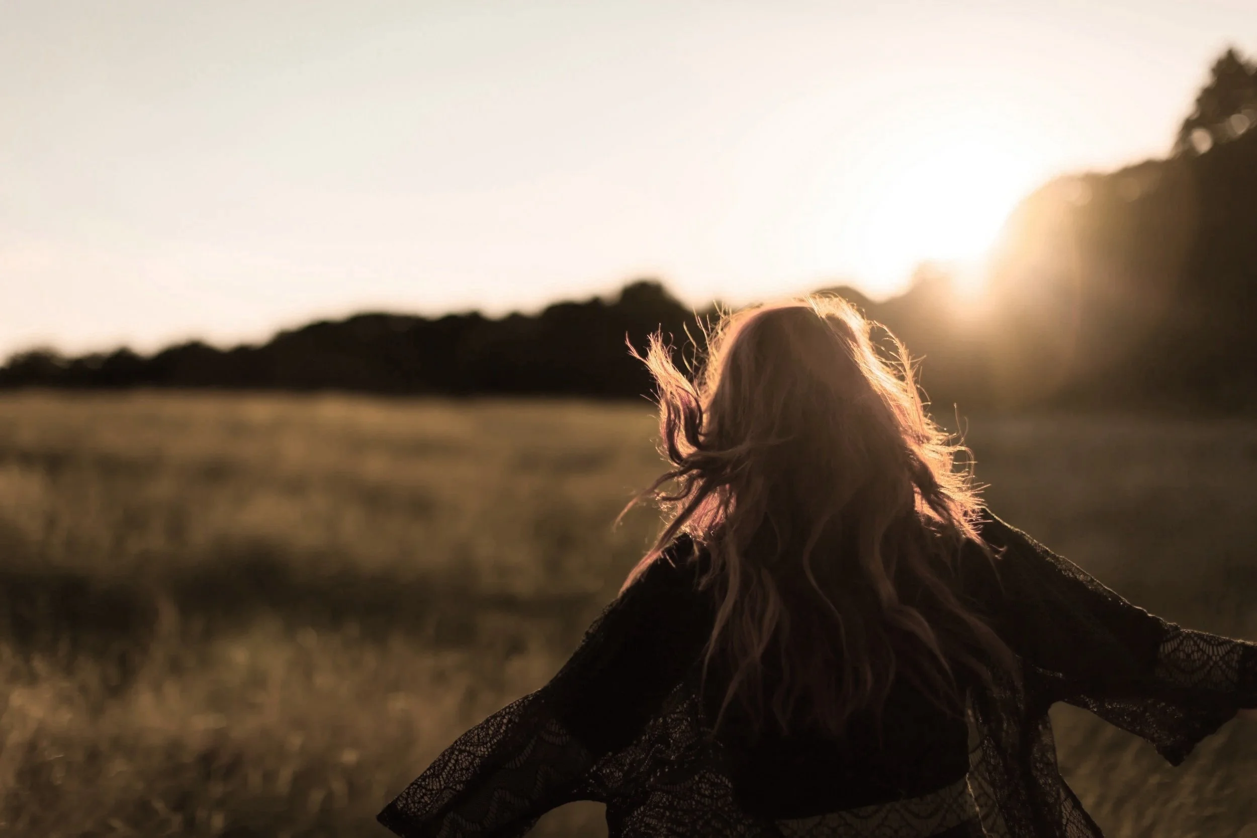 A woman with long, curly hair standing in a field at sunset, facing away from the camera, with her arms outstretched.