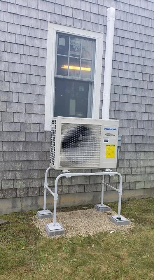 Outdoor air conditioning unit installed with a stand and concrete blocks on grass in front of house wall with window.