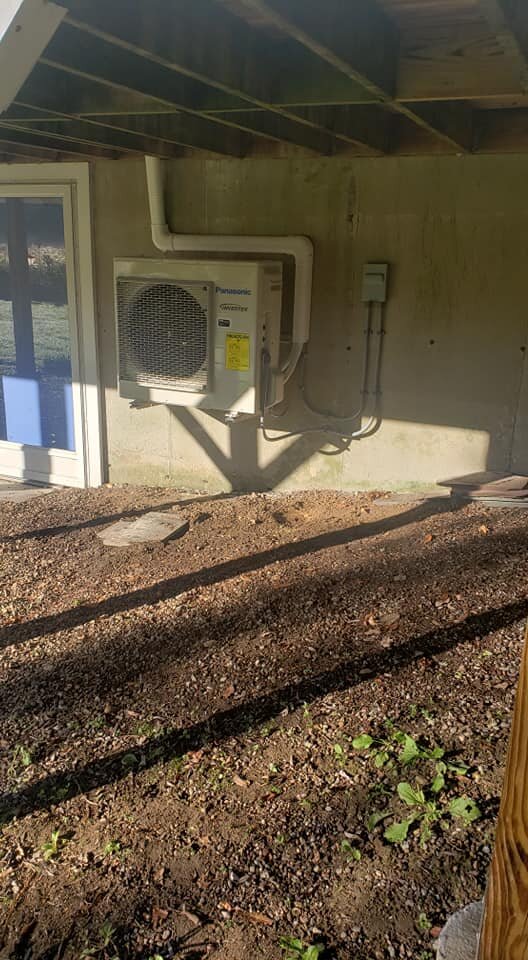 Outdoor view of a building with an air conditioning unit installed on a concrete wall and a sliding glass door, with a dirt ground and shadows cast by nearby structures.