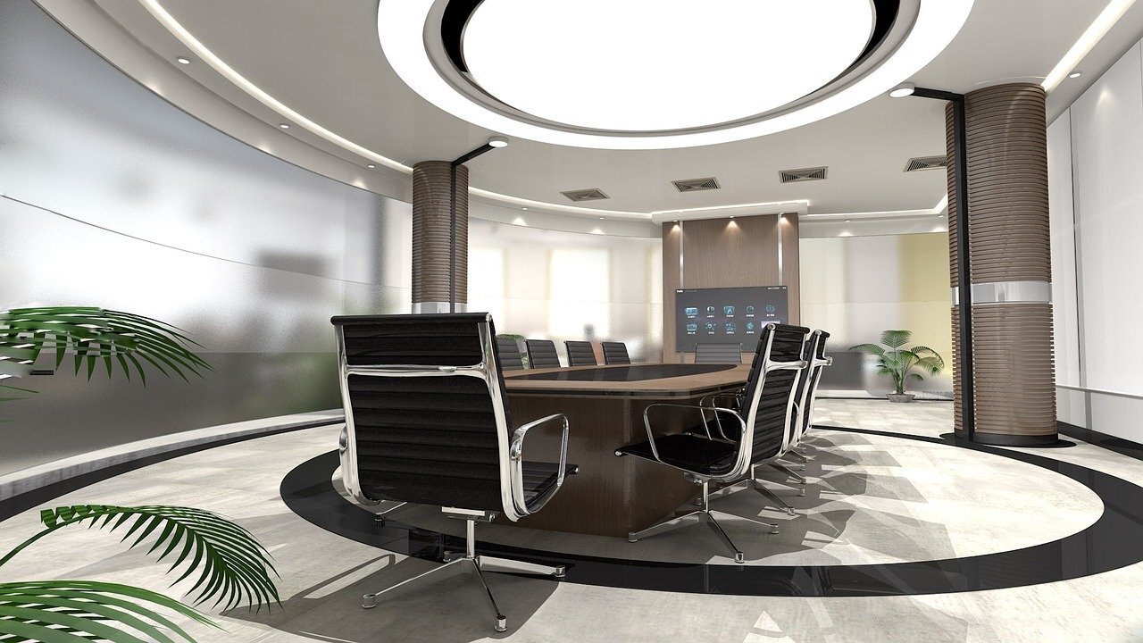 Modern conference room with a large oval table surrounded by black office chairs, and a digital screen on the wall. There are potted plants and large windows with natural light.