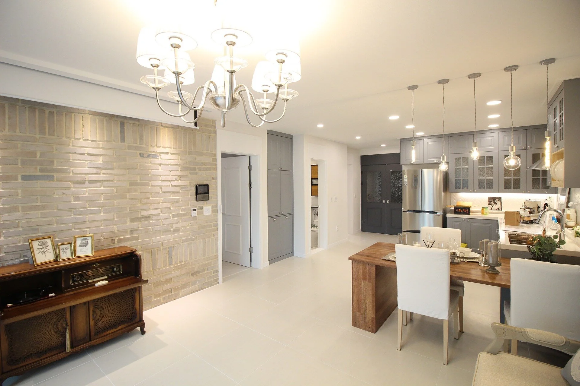 Bright, modern kitchen and dining area with a brick wall, wooden dining table, white chairs, gray cabinetry, stainless steel appliances, and hanging pendant lights.