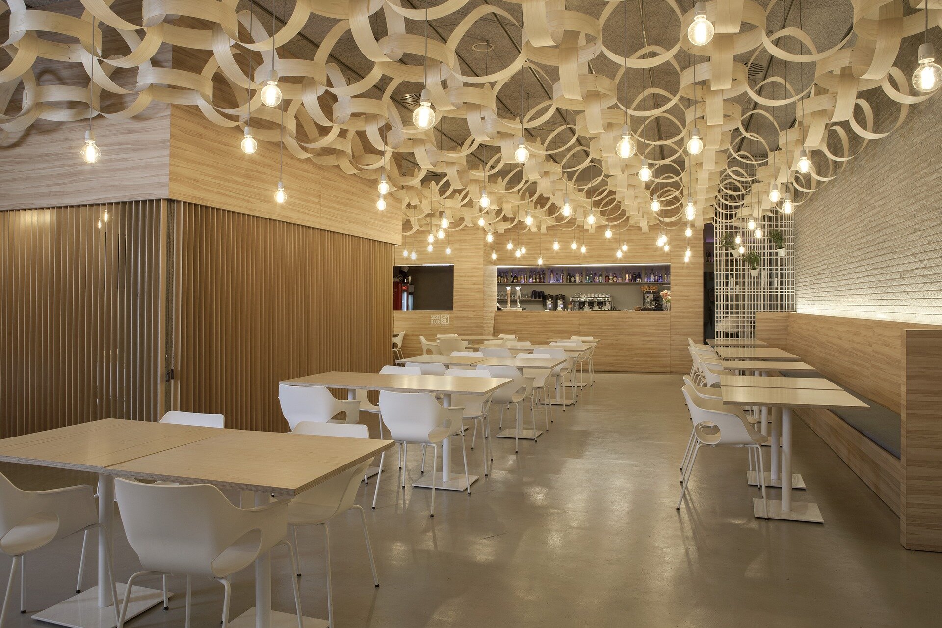 Modern cafe interior with wooden decor, ceiling with hanging light bulbs, white chairs, and tables, and a bar area in the background.