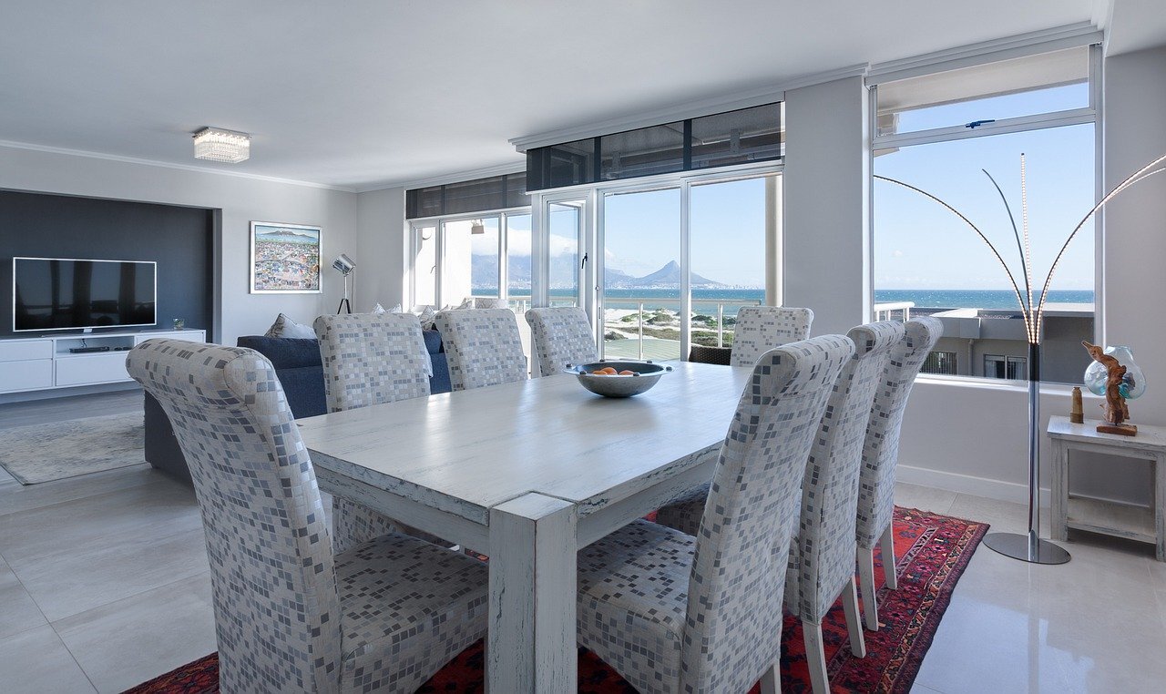 Living and dining area with large windows showing ocean view, white table with patterned chairs, flat-screen TV, and modern decor.