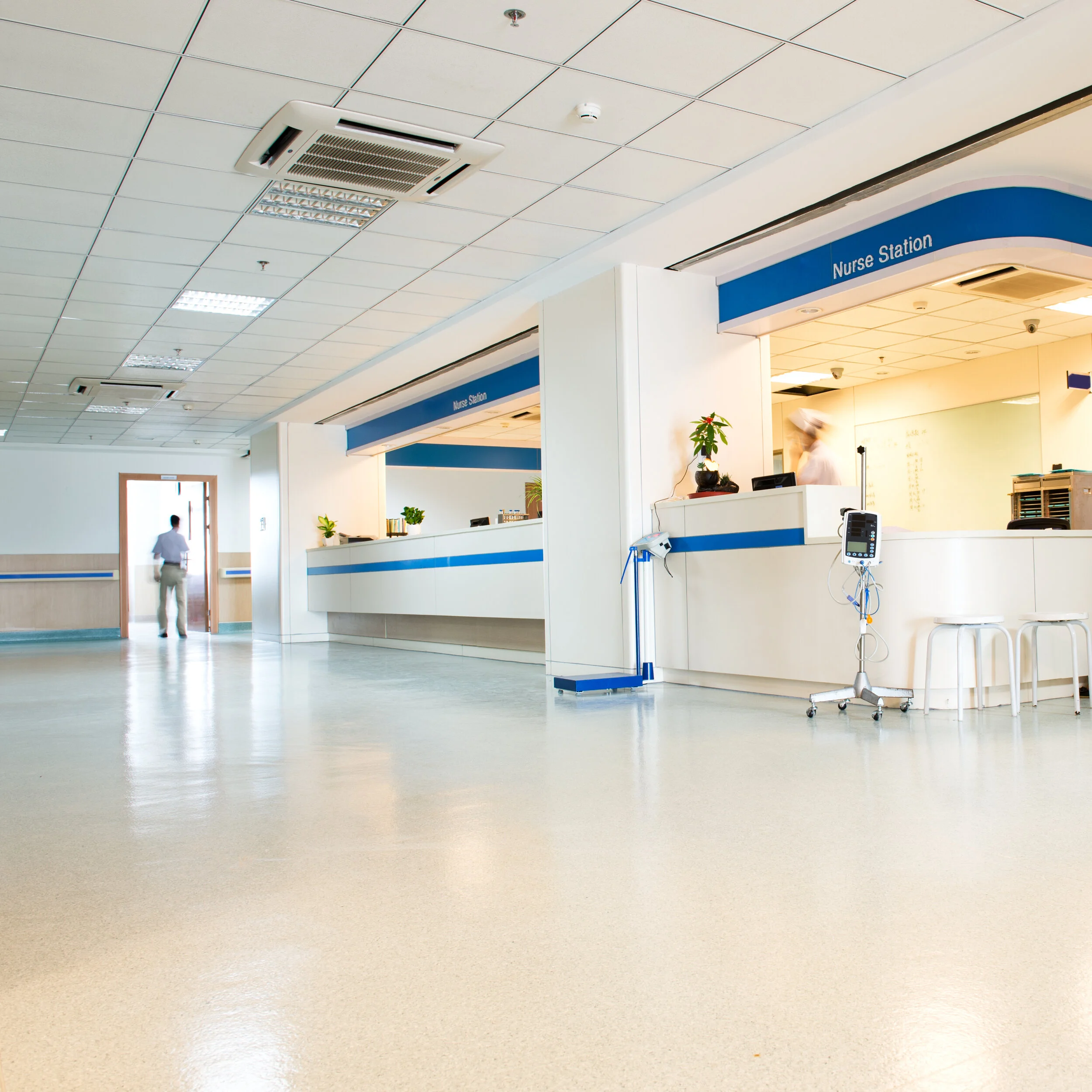 Empty hospital nurse station with a man walking in the background near an open door, medical equipment, and plants.