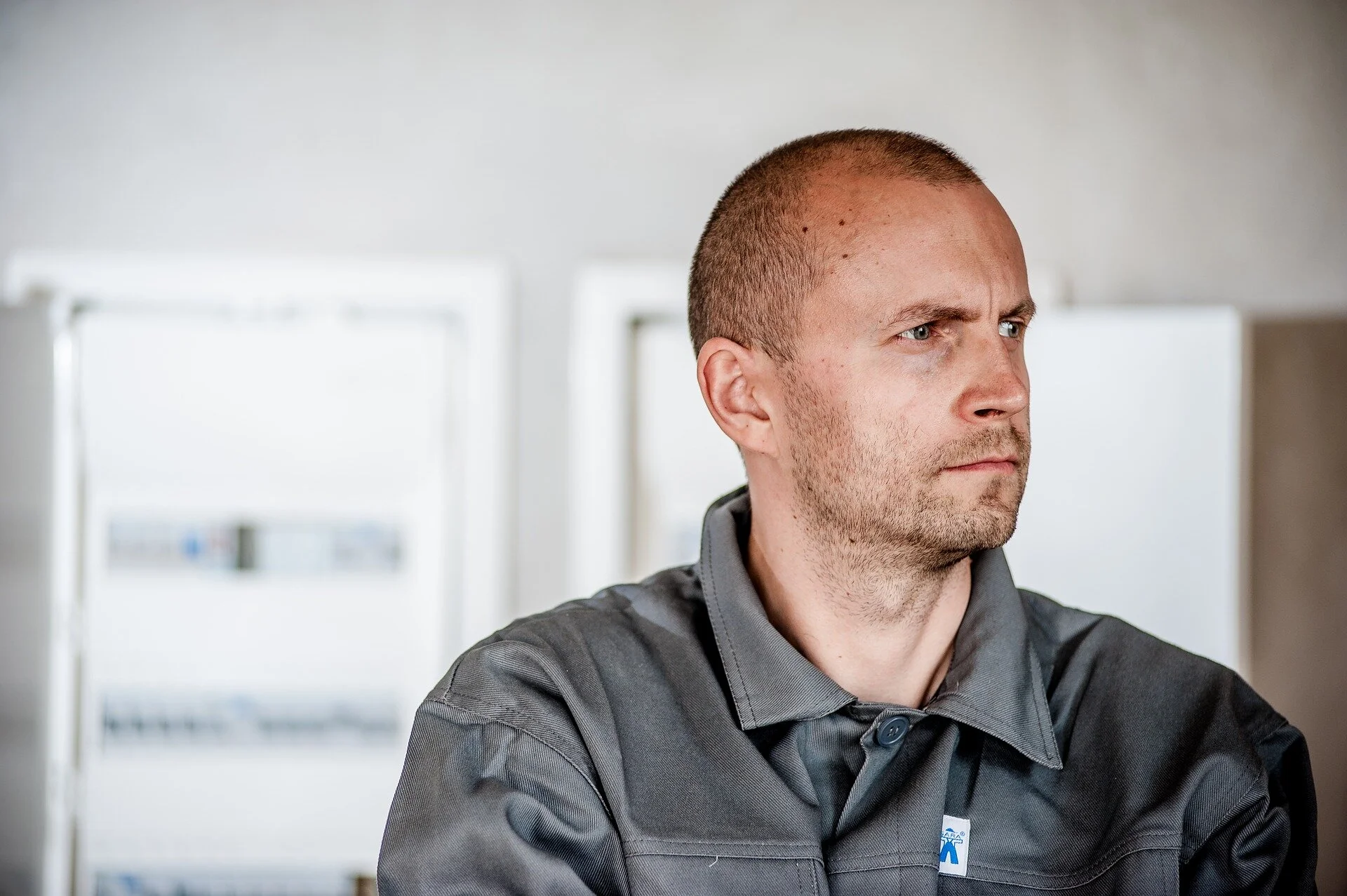 A man with a shaved head and a beard, wearing a gray uniform, looking to the right. The background shows white windows.