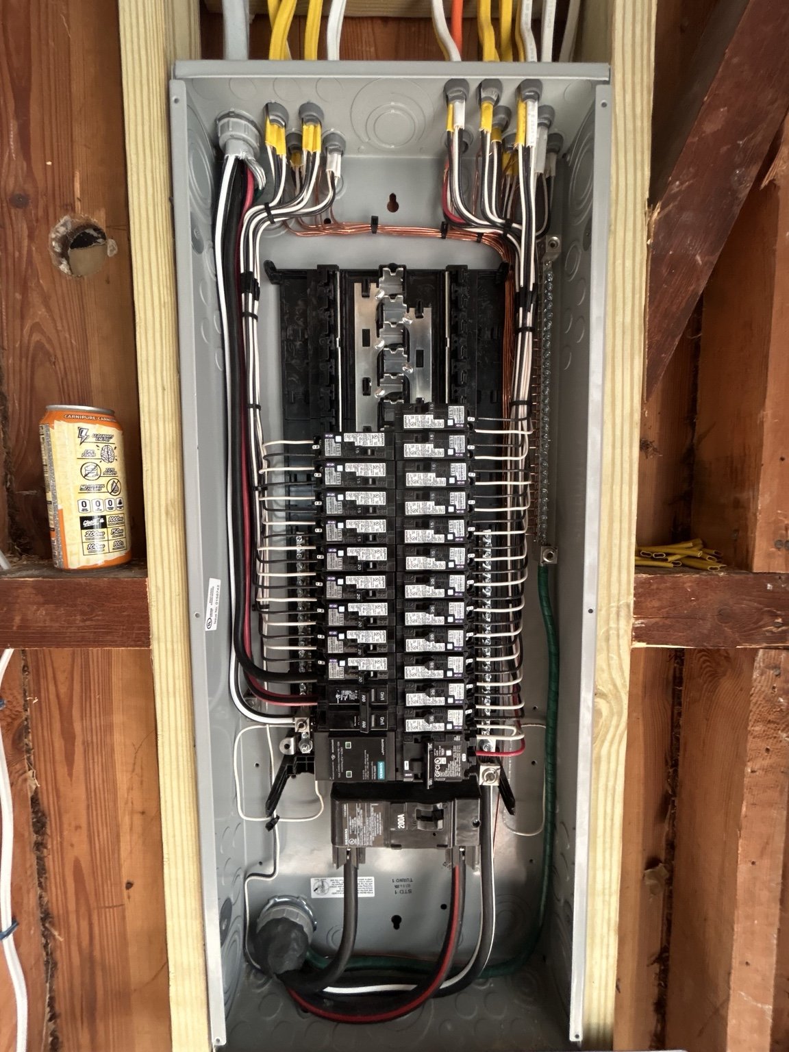 A grey electrical panel with multiple circuit breakers and yellow wiring inside, placed on a workbench with tools and boxes in the background.