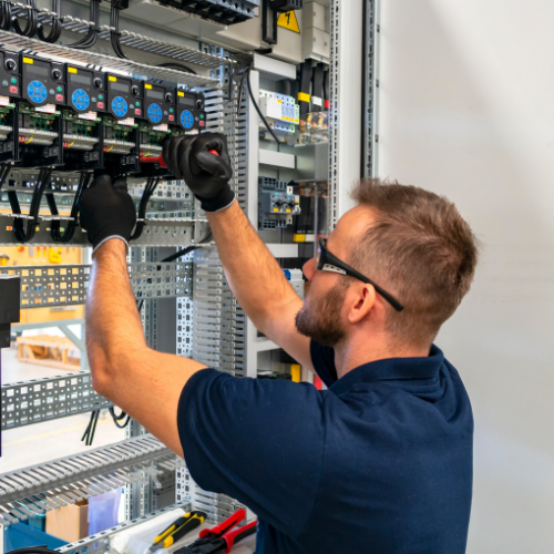 A technician wearing black gloves and glasses working inside an electrical control panel, handling wiring and components.