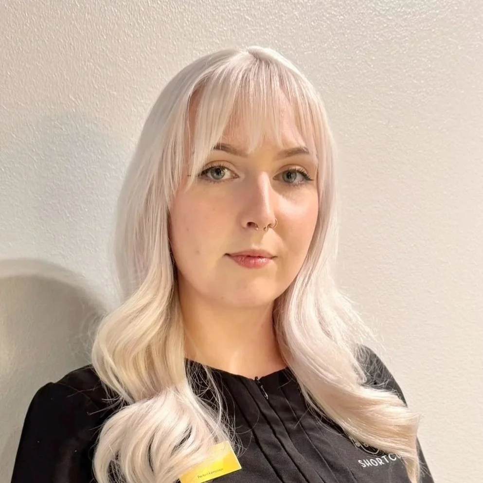 Kuvassa nuori nainen, jolla on vaaleat hiukset, meikki ja korvakorut, seisoo sisätiloissa. Hänellä on päällään musta työpaita, jossa lukee 'Shortcut'. Parturi-kampaaja Krista, Shortcut AALTO