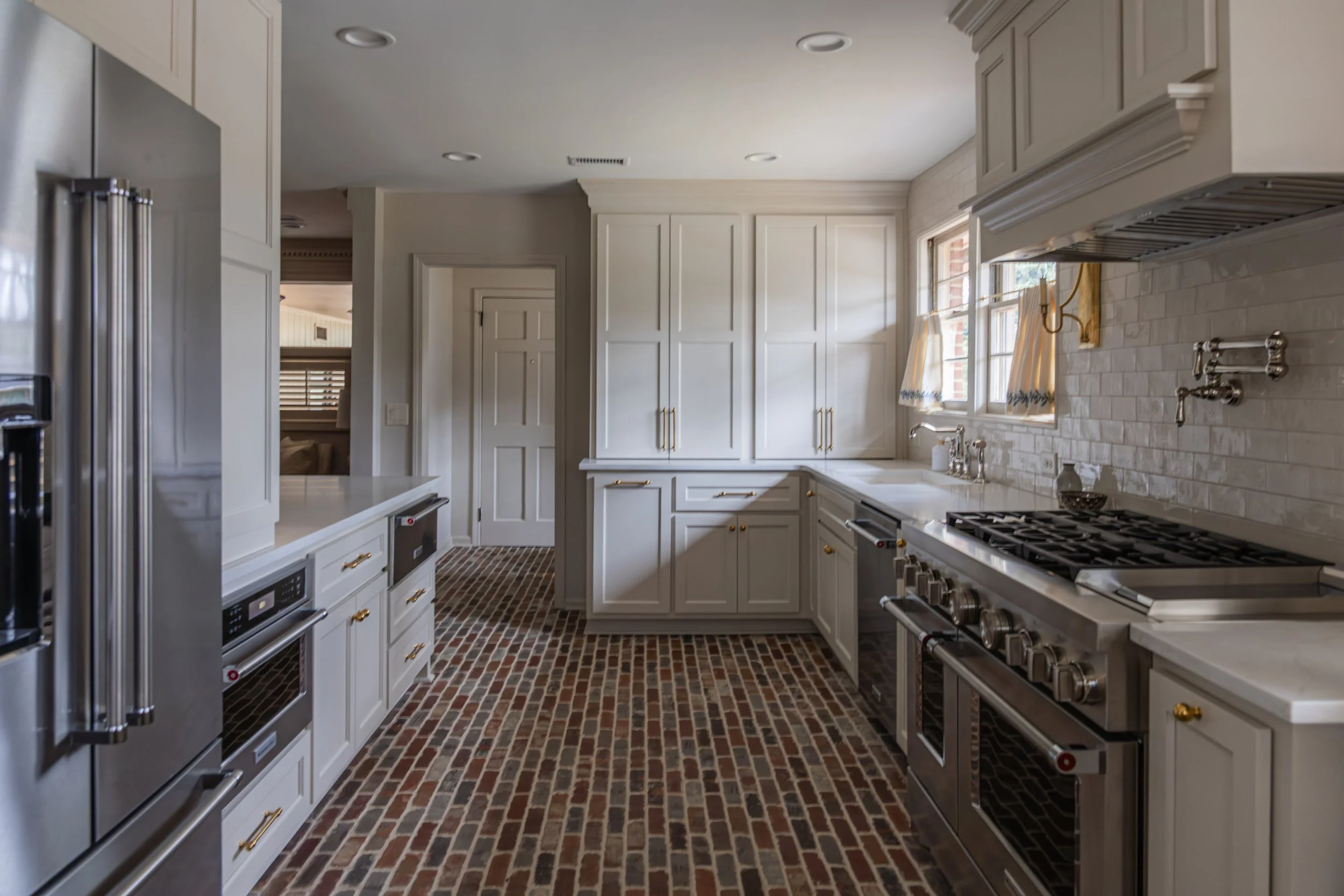 Long galley-style kitchen with white cabinetry, stainless steel refrigerator and range, white tile backsplash, and brick flooring extending through the space.