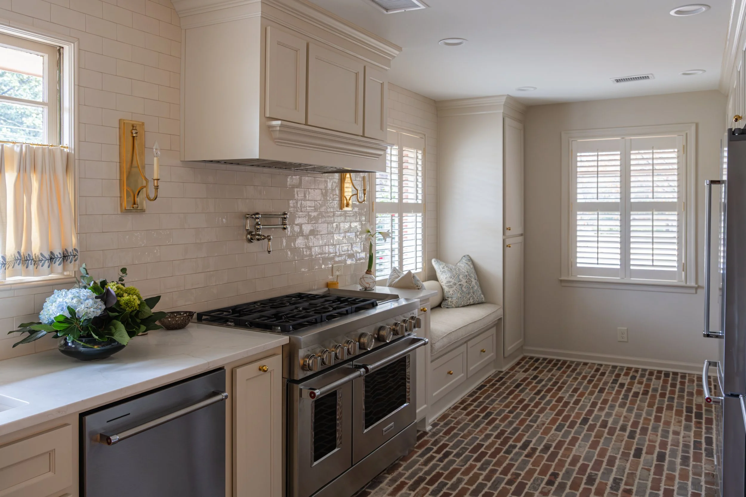 Kitchen corner with stainless steel gas range, white cabinets, white subway tile backsplash, brick flooring, and a windowed breakfast nook with bench seating.