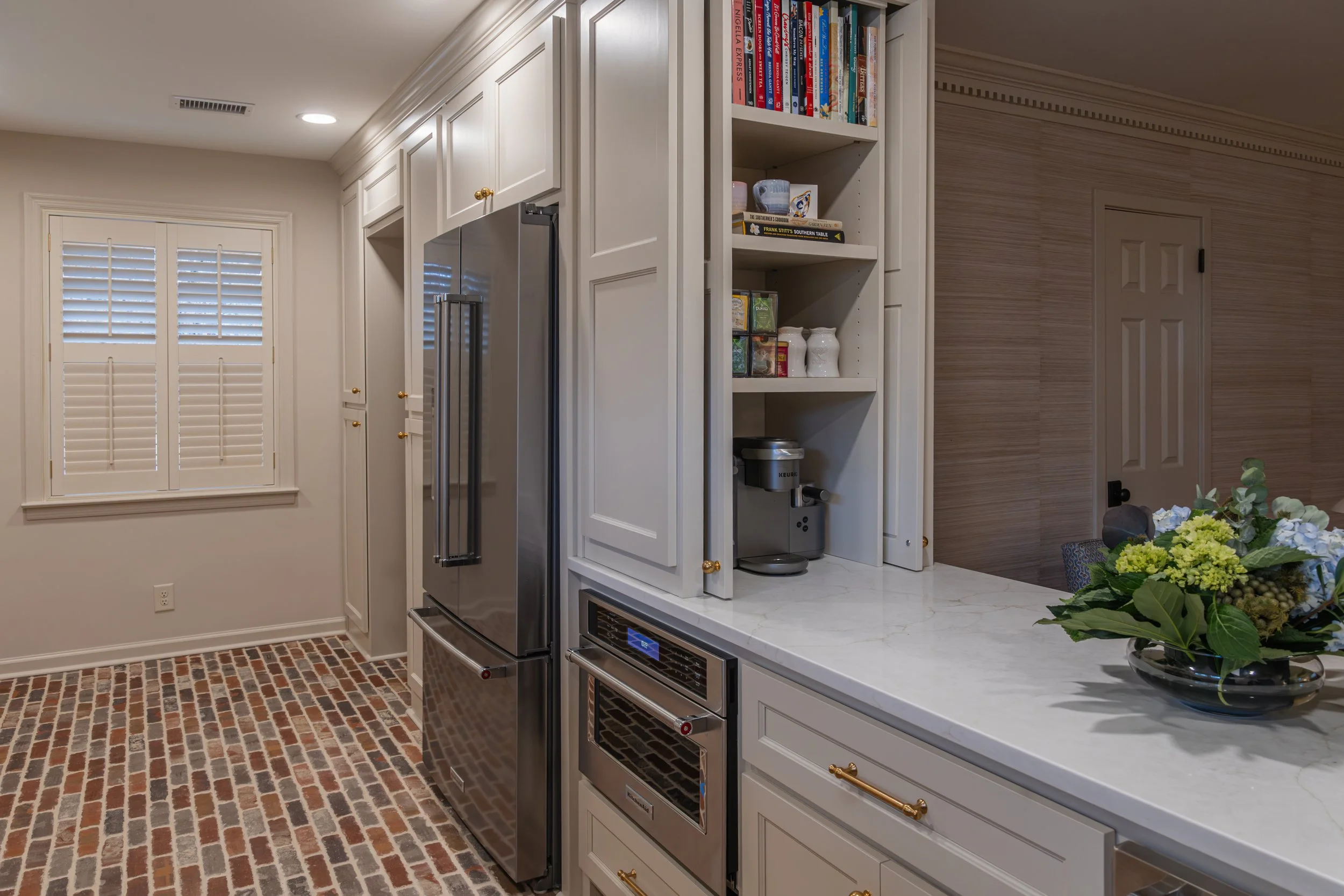 Kitchen wall with built-in cabinetry, stainless steel refrigerator, white countertops, open shelving with books, and brick flooring.