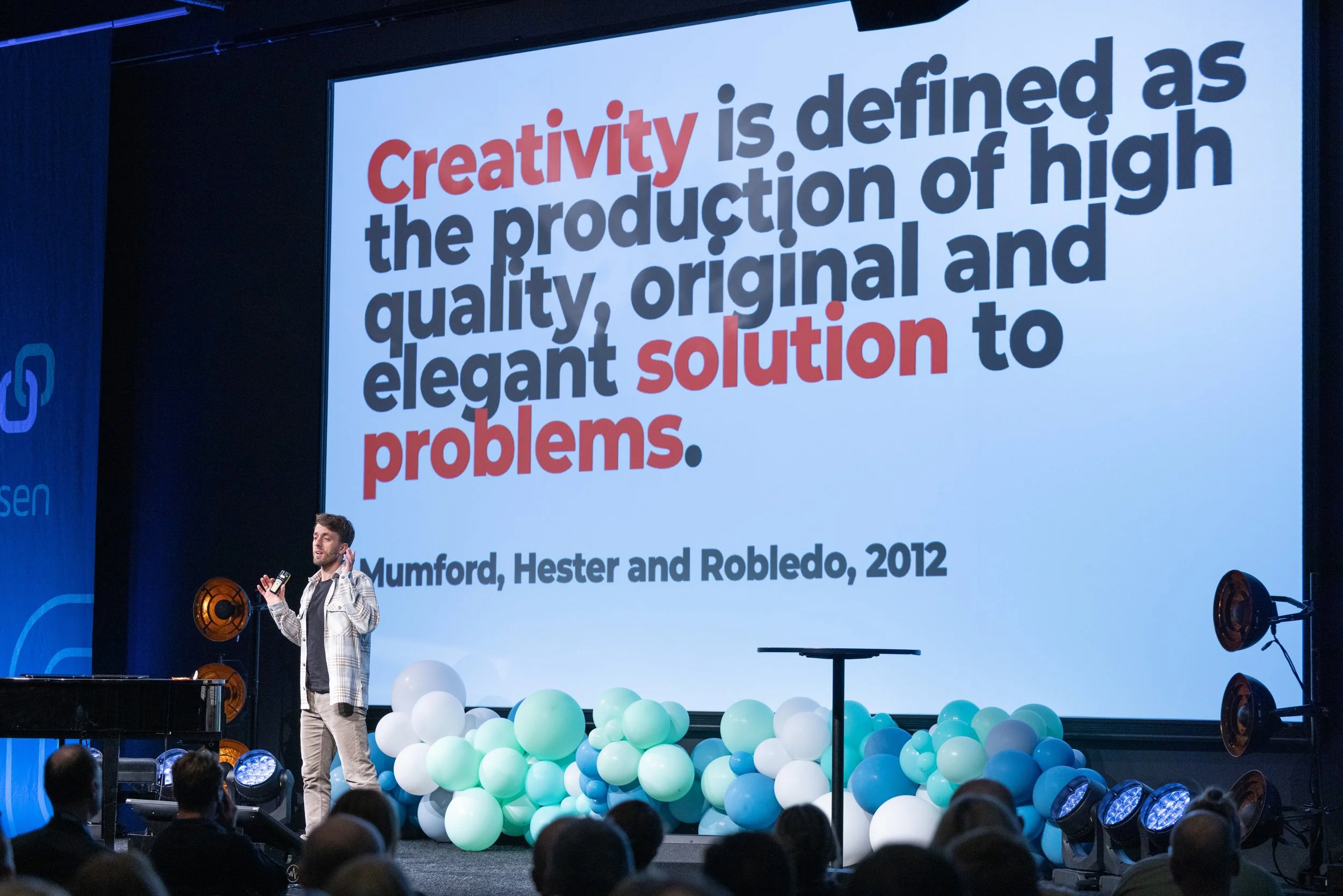 A man giving a presentation on a stage with a large screen behind him displaying a quote. The quote reads, 'Creativity is defined as the production of high quality, original and elegant solutions to problems.' The quote is attributed to Mumford, Hester, and Robledo, 2012. The stage is decorated with blue and white balloons, and there are audience members watching.