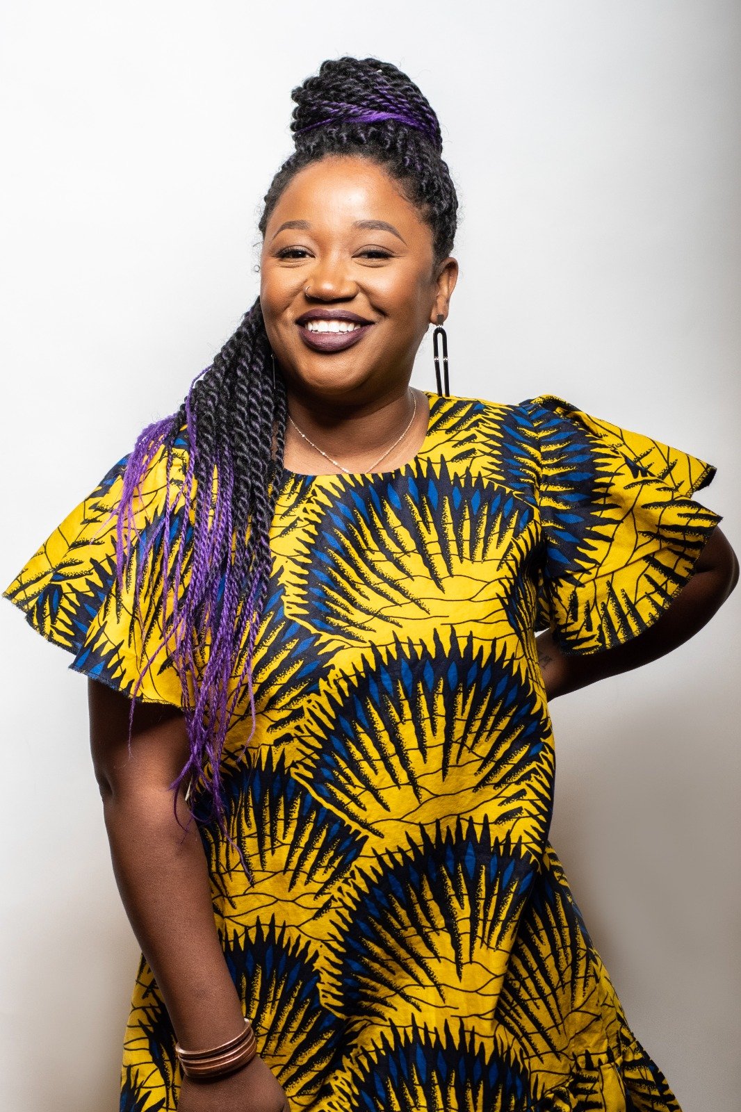 A smiling woman with dark braided hair streaked with purple, wearing a yellow and blue patterned dress, standing against a plain light background.