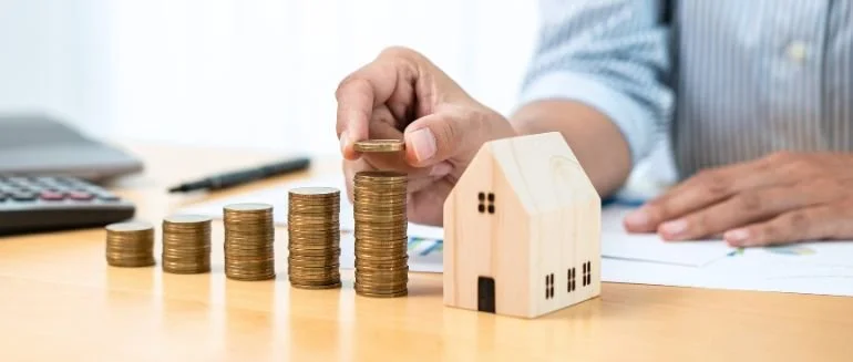 Person stacking coins next to a small wooden house model on a desk, with a calculator and papers nearby.