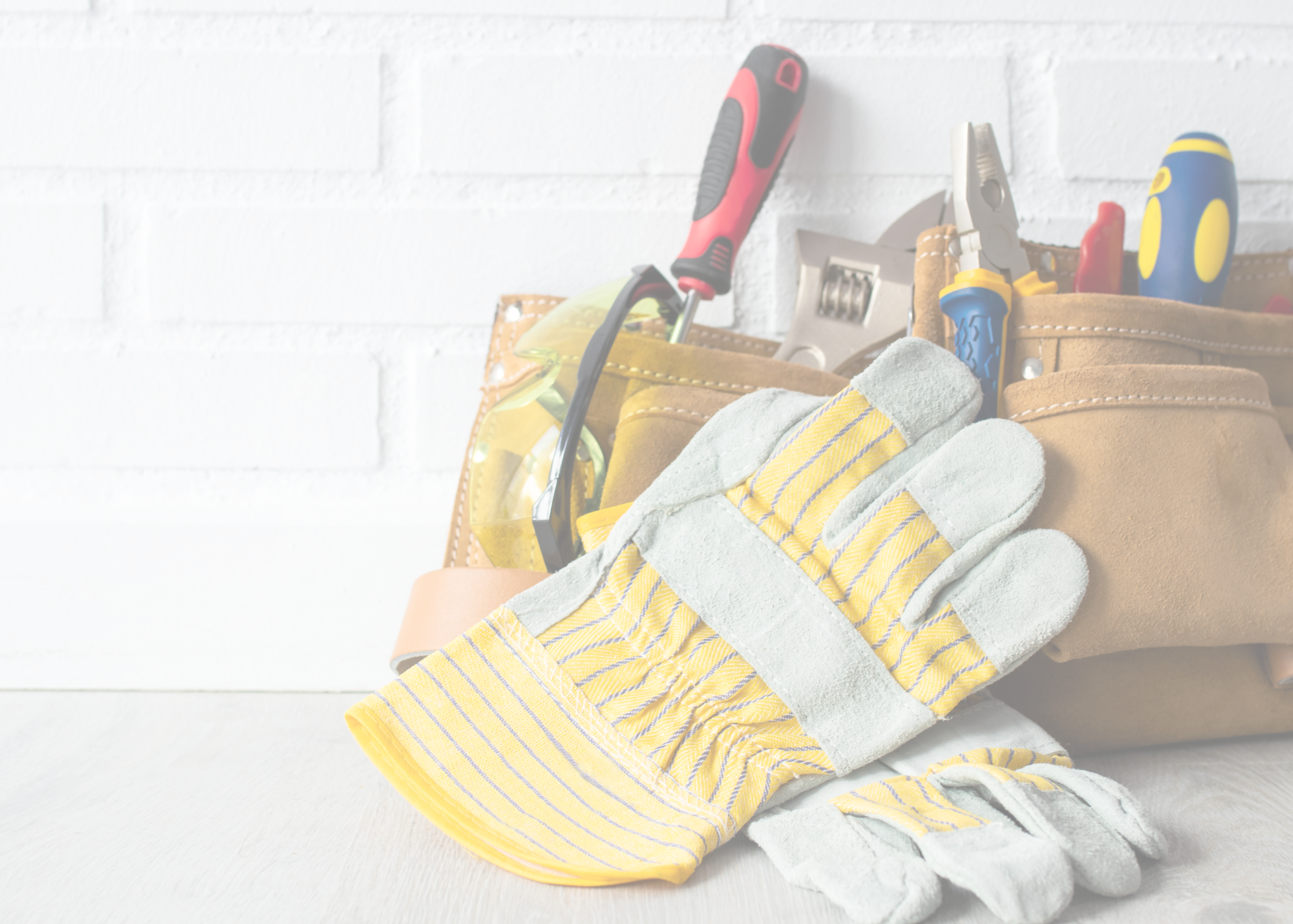 Tool belt on a light wooden surface with work gloves, safety glasses, and various hand tools including screwdrivers, pliers, and a wrench.