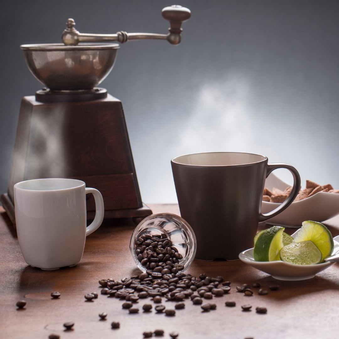 Coffee grinder with coffee beans spilling onto a wooden table, two coffee cups, lime slices, and a bowl of cookies.