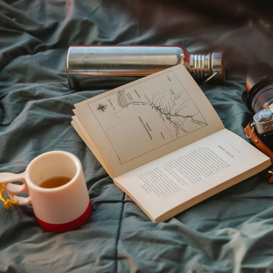 Open book showing a map of the Arkansas River and Navajo Indian Reservation, a cup of tea, a metal water bottle, a camera, and a pair of glasses on a wrinkled gray bedspread.