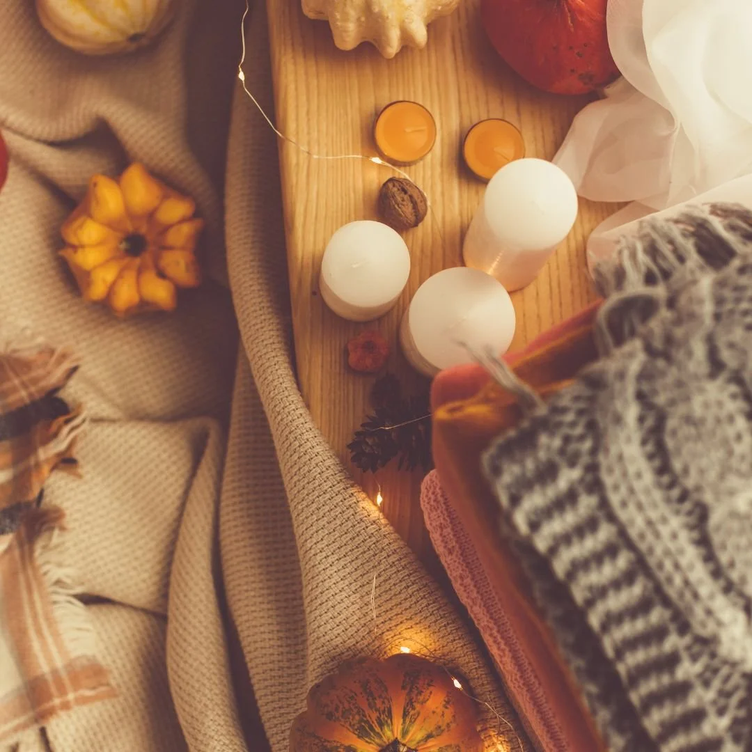 A cozy autumn scene with candles, pumpkins, and warm blankets on a wooden table and fabric, decorated with fairy lights and autumnal decorations.
