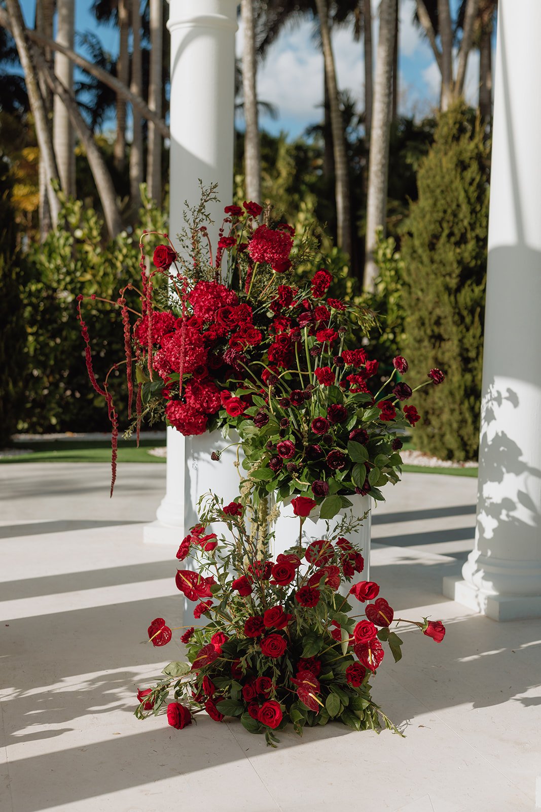 Luxury outdoor wedding ceremony featuring elegant red floral pedestal arrangements beneath a white gazebo, designed by Atico Floral, a Miami and South Florida wedding florist.