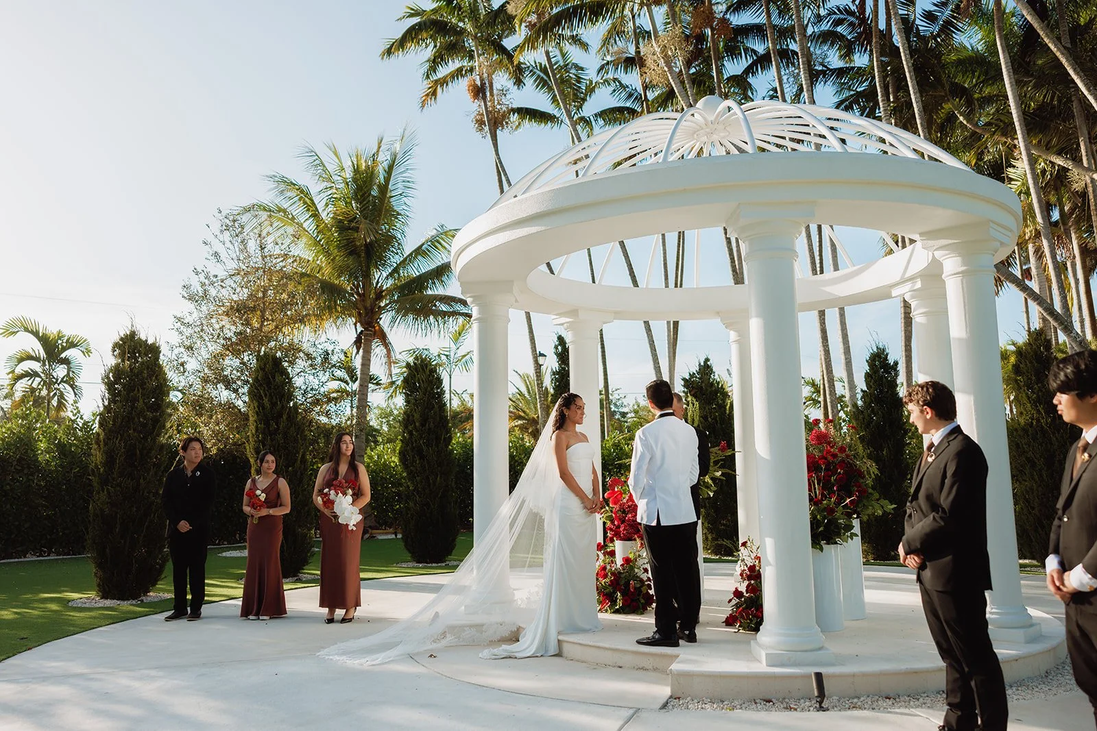Looking for luxury wedding ceremony inspiration? This elegant white gazebo is adorned with lush red floral arrangements featuring premium roses, orchids, and cascading accents to create a romantic outdoor wedding ceremony in South Florida.