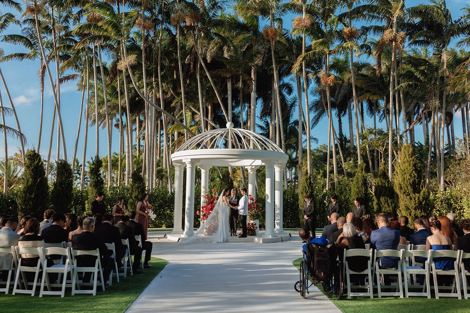 Looking for luxury wedding ceremony inspiration? This elegant white gazebo is adorned with lush red floral arrangements featuring premium roses, orchids, and cascading accents to create a romantic outdoor wedding ceremony in South Florida.